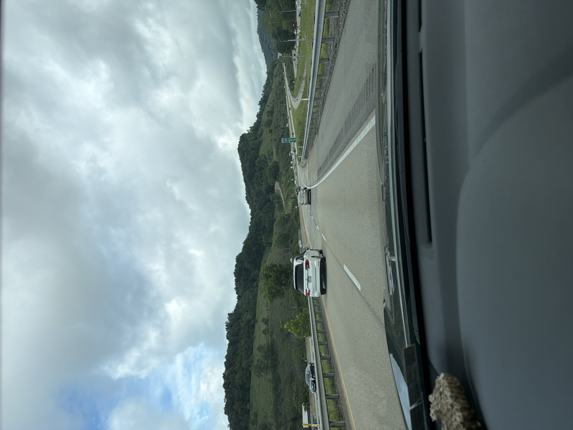 The image captures a view from inside a vehicle driving on a highway in Ohio Township, United States of America. The highway appears to be multi-lane, with other vehicles visible on the road. The surrounding environment consists of rolling green hills covered in trees, suggesting a semi-rural or suburban setting. The sky is overcast with a mix of clouds and patches of blue sky. A guardrail separates the lanes of traffic and borders the shoulder. Road signs are visible, indicating direction and distance. The dashboard of the vehicle is partially visible in the foreground, with a small object on the left side.