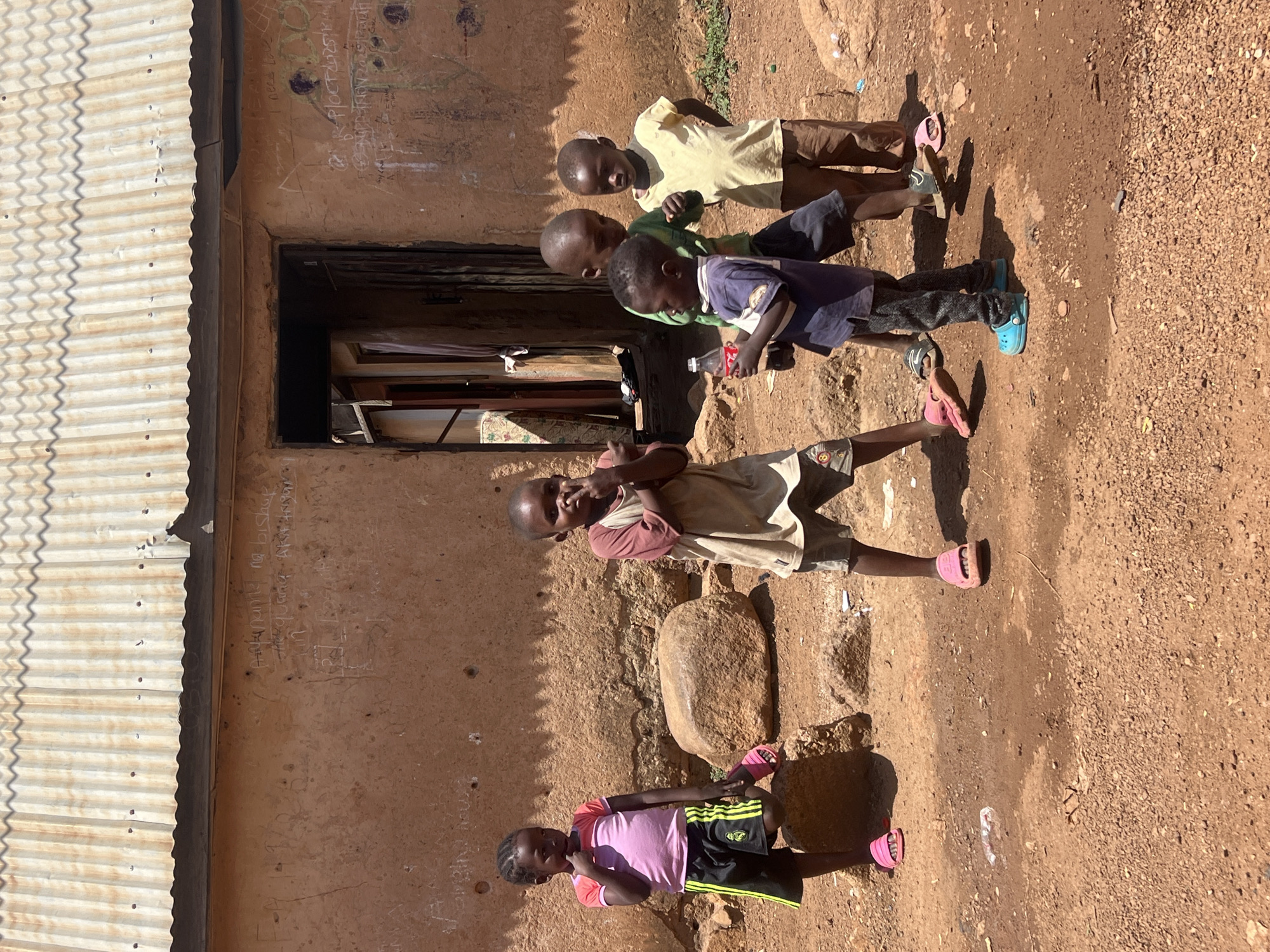 Five children stand outside a building with a corrugated metal roof in Gura Top, Nigeria. The building's walls are a faded reddish-brown, and graffiti is visible. The ground is dirt and gravel. The children are wearing varied clothing, including t-shirts, shorts, and sandals, many of which are pink. One child holds a small, red plastic bottle. The children appear to be in a sunny outdoor setting.