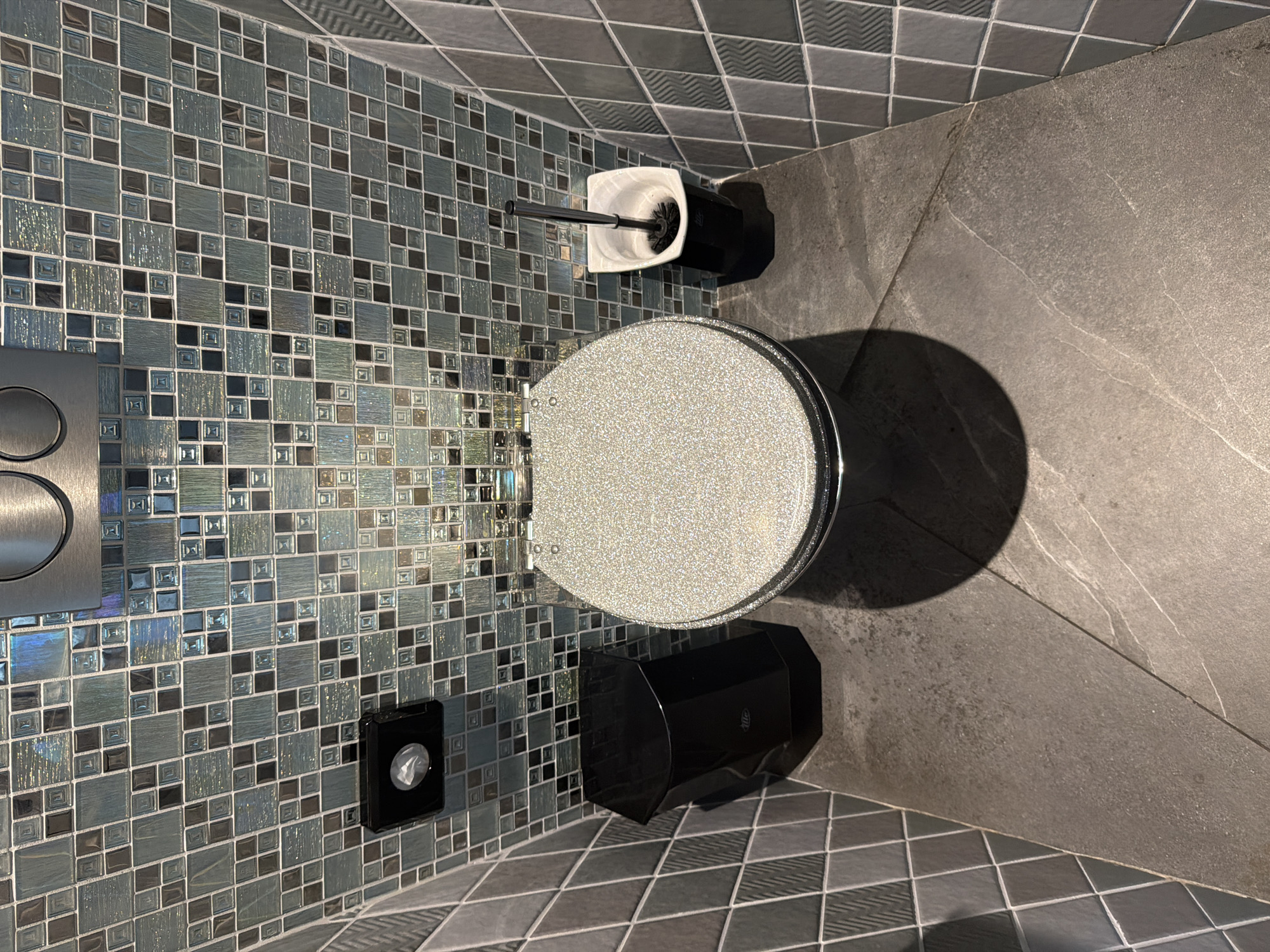 The image shows a top-down view of a modern toilet in a bathroom, possibly located in Amstelveen, Netherlands. The toilet is white with a speckled, shimmering seat and lid. To the left of the toilet is a black waste bin. To the right is a white toilet brush holder with a black toilet brush. The walls are covered in a mosaic of small, iridescent square tiles in shades of blue, green, and gray. The floor appears to be made of large, dark gray tiles with a subtle veining pattern. A metal flush plate is visible at the top of the image, mounted on the tiled wall above the toilet. The lighting creates a shadow of the toilet on the floor. There are no people or animals in the image.