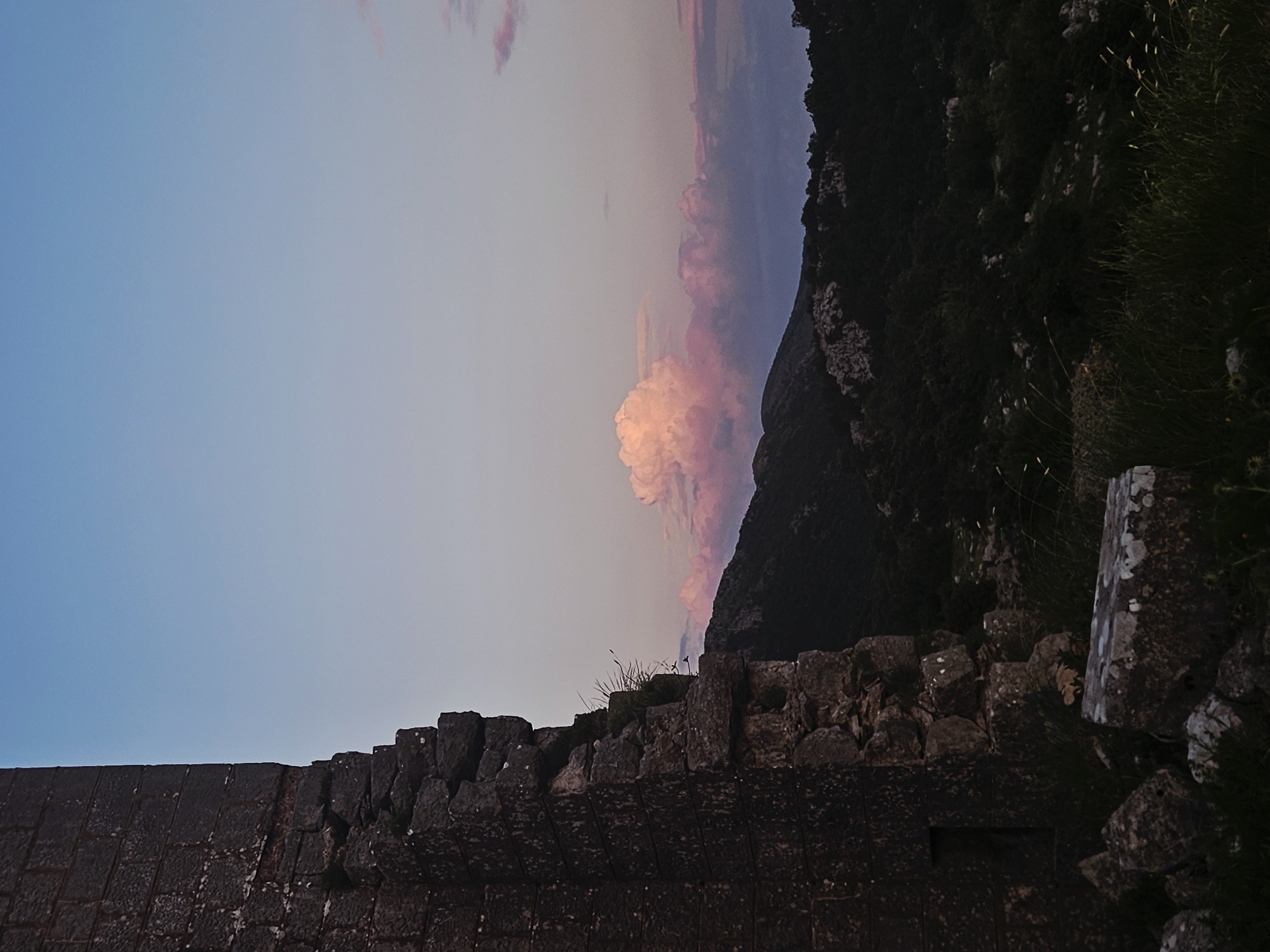 The image shows a landscape in Unknown, Unknown, featuring a combination of natural and man-made elements under a sky with pink-tinged clouds. In the foreground, there's a stone structure, likely a wall or ruin, constructed from dark, irregular stones. Some vegetation grows on top of this structure. Behind the wall, a mountain rises steeply, covered in dense, dark green vegetation. The sky above is a mix of blues and purples, with a prominent pink cloud formation. The overall scene has a tranquil, naturalistic feel.