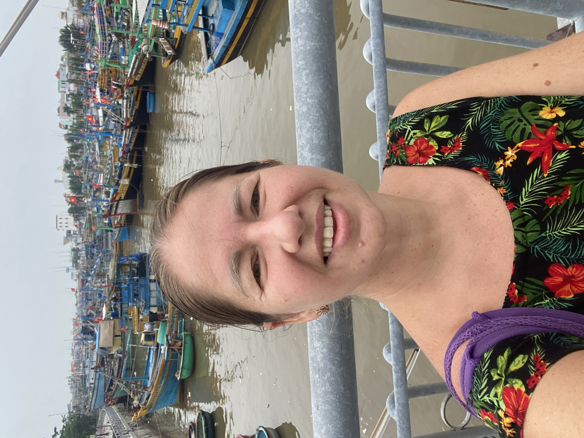 In the city of Phan Thiết, Vietnam, a woman with shoulder-length brown hair takes a selfie on a bridge overlooking a harbor filled with numerous fishing boats. The boats are of various sizes and colors, predominantly blue, yellow, and green, with some displaying red flags. The water in the harbor is a murky brown. In the background, buildings and trees are visible along the shoreline. The woman is wearing a black sleeveless dress with a floral pattern of red hibiscus flowers and green leaves. A purple strap from a bag is visible over her left shoulder. She is smiling at the camera, and her face is well-lit. The railing of the bridge she is leaning on is made of grey metal. The sky is overcast.