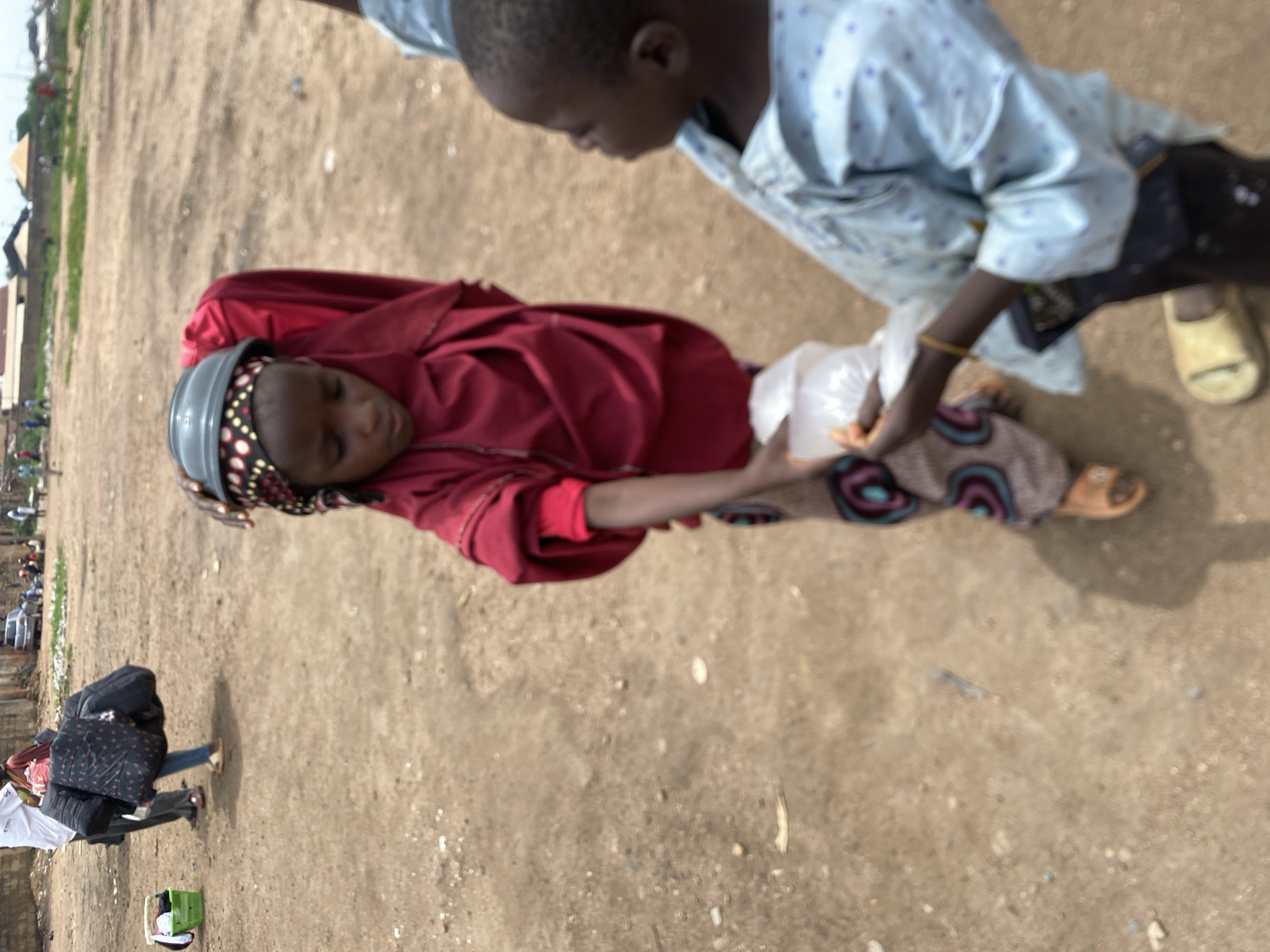 In Bukuru, Nigeria, two children are the primary subjects. One child, wearing a red garment and patterned head covering, appears to be receiving a plastic bag from another child. The second child is dressed in a light blue patterned shirt and dark shorts. They stand on a sandy ground. In the background, there are buildings, vehicles, and other people visible. A woman carrying a large bag walks in the lower-left corner of the image.
