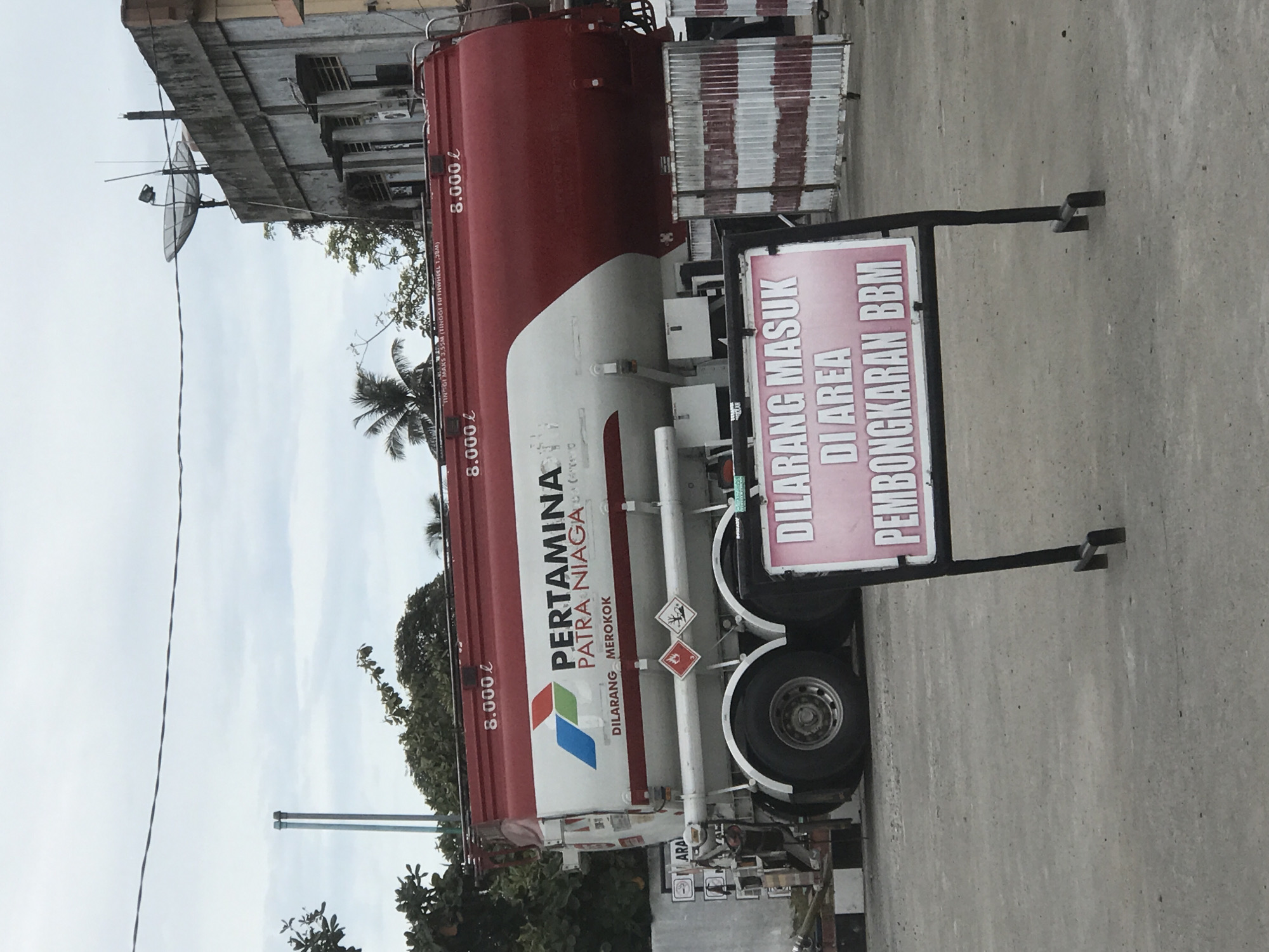 The image shows a Pertamina Patra Niaga fuel tanker truck parked on a concrete surface. A sign is standing beside it that reads "DILARANG MASUK DI AREA PEMBONGKARAN BBM" (meaning "No entry to the fuel unloading area" in Indonesian). The tanker is red and white with the Pertamina logo and the phrase "DILARANG MEROKOK" ("No Smoking"). The text "8.000 l" is written on the tanker three times. Behind the truck, there is a building with a satellite dish attached. There are trees and other vegetation visible as well. The sky is overcast.