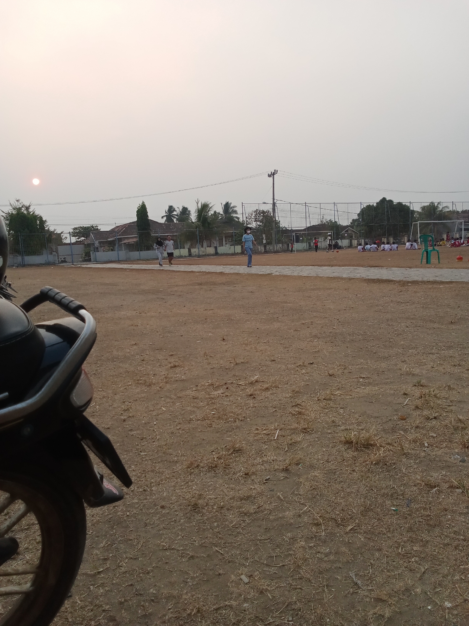 The image captures a dusty, outdoor scene, possibly in Toto Katon, Indonesia, during what appears to be late afternoon or early morning due to the soft, diffused sunlight. In the foreground, the rear portion of a dark-colored motorcycle is visible on the left, with its tire, fender, and a portion of the seat. The ground is dry and covered with sparse, light-brown vegetation and dirt.
In the mid-ground, a fenced-off sports field, likely a soccer field, is present. Several people are visible on or near the field. A person wearing a blue shirt and a mask walks on the left side of the field. Further into the field, two individuals are running, one in white and the other in dark clothing. In the background, a group of children in white uniforms are seated on the ground, facing a soccer goal. Another group of children in red uniforms are also present further to the right. A green plastic chair stands near the seated children. In the far background, houses with tiled roofs and palm trees are visible, suggesting a residential area surrounding the field. The sky is overcast with a hazy, light-colored tone, and the sun appears as a faint, bright circle in the upper left quadrant.