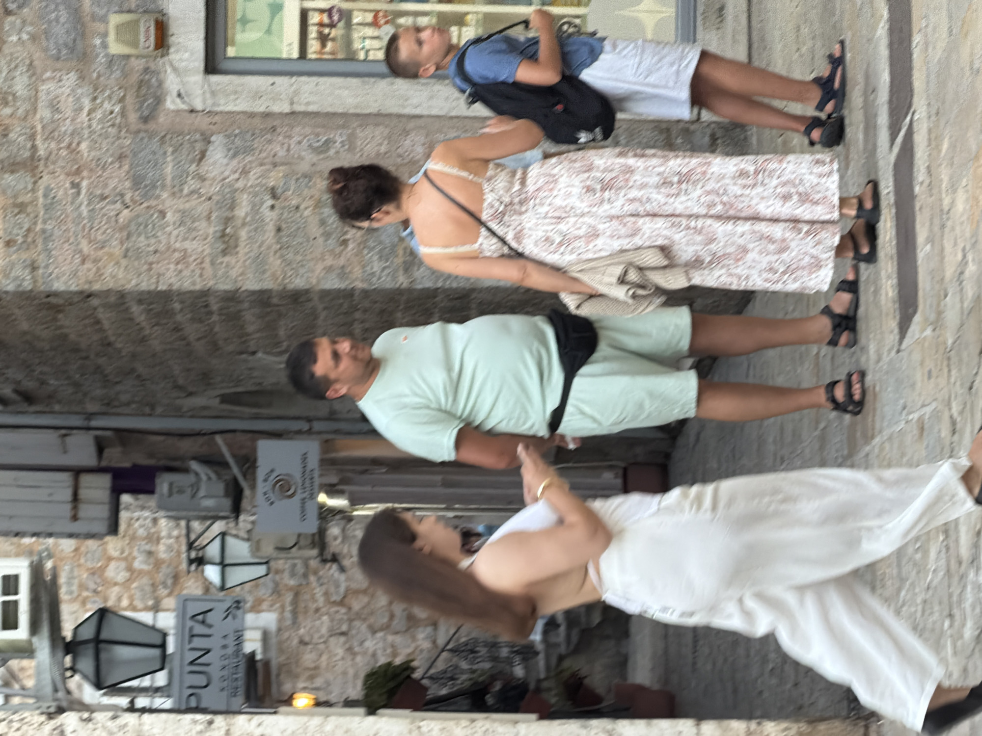 In Budva, Montenegro, a group of four people are walking on a cobblestone street. A woman in the foreground walks from left to right, wearing loose white pants and a white top. She is looking at her phone, which she holds in her right hand. To her right stands a man in a light green t-shirt and shorts, with a black fanny pack around his waist. He is looking forward. Behind him, a woman in a white and pink floral dress walks to the right. She carries a folded tan garment over her left arm and has a black strap across her chest. A young boy in a blue t-shirt and white shorts walks to the right behind the woman in the floral dress, carrying a black backpack. The background shows stone buildings with lanterns and signs. One sign, partially visible, reads "PUNTA RESTAURANT." The scene appears to be during the day, with soft lighting.