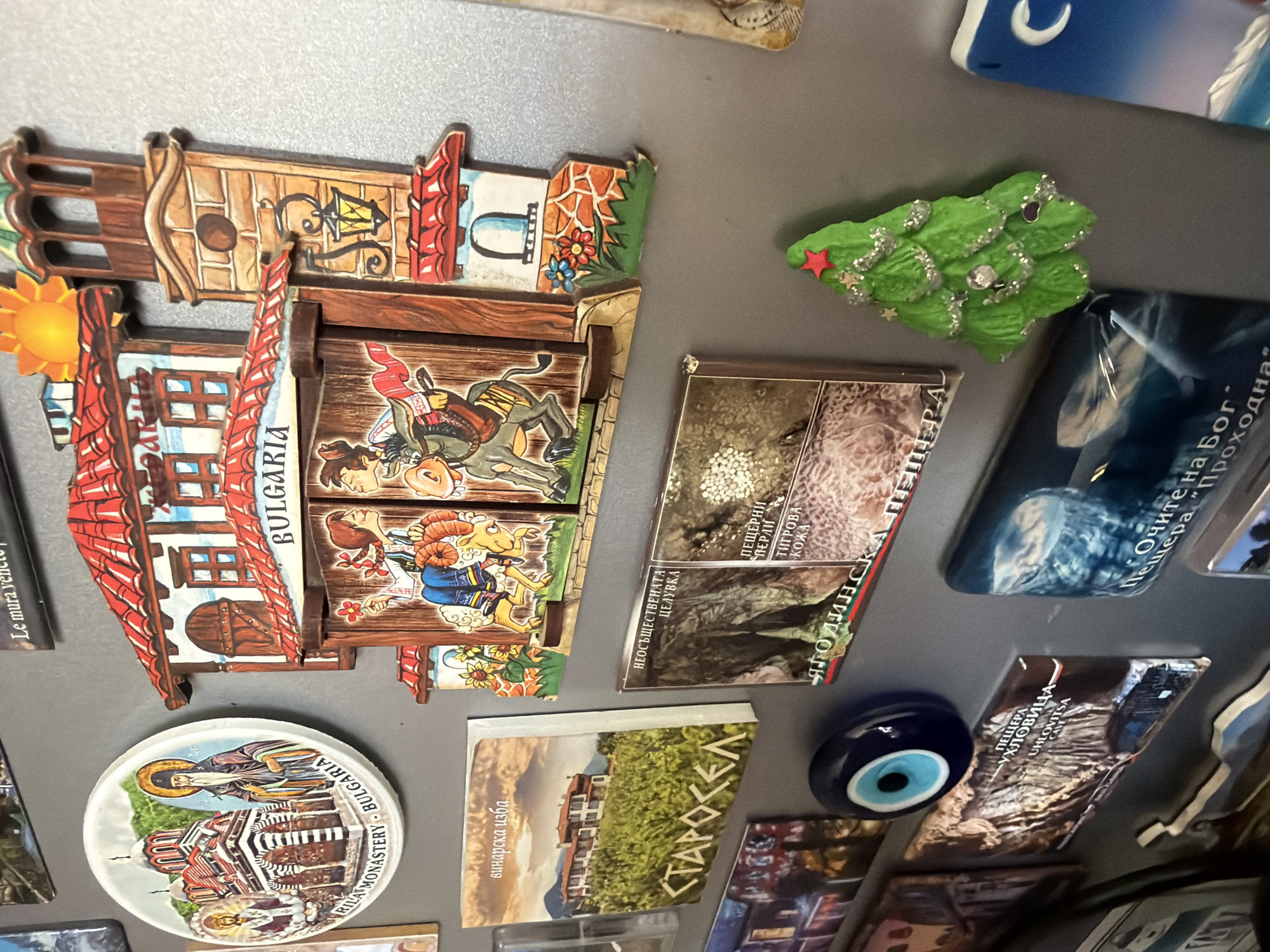The image displays a collection of magnets on a gray, metallic surface, likely a refrigerator door, located in Pazardzhik, Bulgaria. The magnets feature various designs, including a circular magnet depicting Rila Monastery with a figure of a monk, a multi-dimensional magnet with a traditional Bulgarian house facade and figures of people and a donkey, and rectangular magnets with images of Bulgarian landscapes and text in the Bulgarian language. One magnet shows a vineyard with a building labeled "СТАРОСЕЛ", another depicts a cave entrance labeled "УХЛОВИЦА", and a third magnet features a dark, rocky landscape with text. There is also a blue evil eye charm magnet and a green Christmas tree-shaped magnet. The overall impression is a display of souvenirs and decorations related to Bulgarian culture and landmarks.