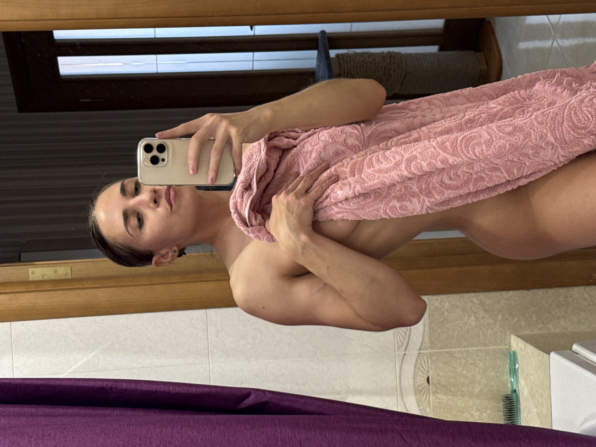 A person is captured in a mirror selfie, holding a light-colored smartphone with three visible rear cameras. The individual, who appears to be female, is partially covered by a light pink towel featuring an intricate, embossed pattern. Their expression is neutral, with a slight pout.
The setting appears to be a bathroom, characterized by a light-colored tiled wall visible in the background and a dark purple fabric, possibly a shower curtain, prominent in the lower foreground. A wooden frame or trim runs horizontally across the upper portion of the scene, potentially part of the mirror's frame or a window/doorway. In the bottom right corner, a toothbrush with a light blue handle is faintly visible on a counter or ledge. The person's hair appears somewhat damp, suggesting a recent shower or bath. No specific time of day or external weather conditions are discernible. There is no visible text in the image.
