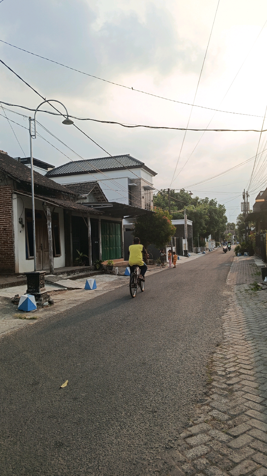 A street scene in Kertosari, Indonesia, at dusk, features a person riding a bicycle down the center of an asphalt road. The cyclist, wearing a yellow shirt, is in motion, with the bicycle's wheels blurred. To the left of the road are several houses with traditional Indonesian architecture, characterized by tiled roofs and open verandas. Decorative, blue and white conical markers are placed along the edge of the sidewalk. To the right of the road, a paved sidewalk made of interlocking bricks runs parallel. Further down the road, a few children are standing on the sidewalk, and a motorcycle is visible in the distance. The sky is overcast with diffused light, indicating either early morning or late afternoon. Power lines crisscross the sky above the street and houses.