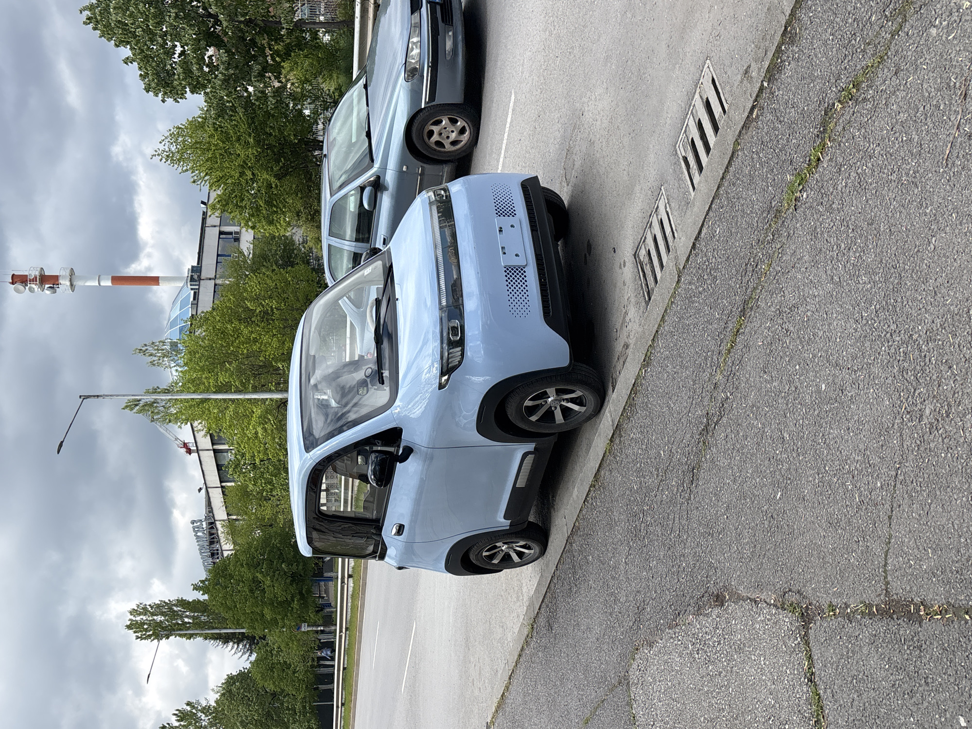 A light blue, compact electric vehicle, resembling a microcar, is parked on an asphalt surface alongside a curb. Its front end, featuring a textured gridded pattern and an empty license plate bracket, faces towards the viewer. Behind it, a grey sedan is also parked, with its front wheel and side visible. To the right, the asphalt surface transitions to a curb with two metal drainage grates embedded in it.
In the background, a dense line of green trees and bushes obscures parts of several buildings. A prominent red and white striped telecommunications tower or chimney, equipped with multiple antennas, stands tall on the left side of the frame. Further right in the background, a large sign with Cyrillic text is partially visible through the trees; the readable portion spells out "БИОХИМ" (BIOCHEM). Streetlights are also discernible in the distance. The sky is overcast with heavy clouds, suggesting a cloudy daytime scene. The overall setting appears to be an urban or industrial area in Sofia, Bulgaria.