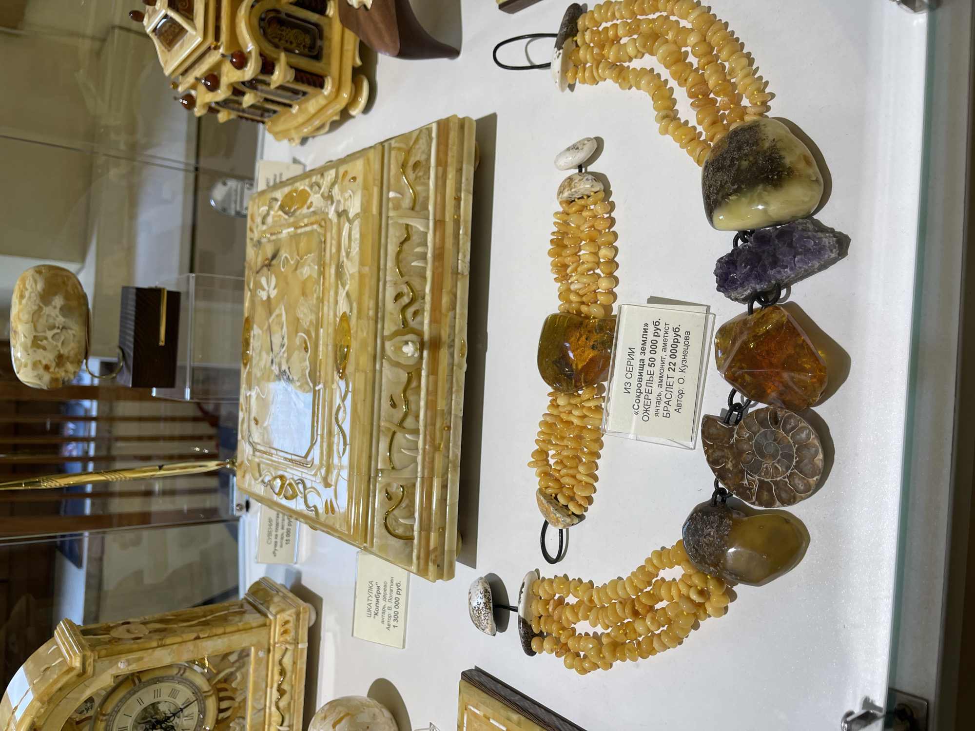 The image shows a collection of amber artifacts displayed in a glass case, likely in a museum or gallery. A square amber box with ornate carvings is prominently featured, along with an amber clock and several other amber decorative items. To the right, there is a necklace and bracelet made of amber beads and semi-precious stones like amethyst and fossilized ammonite. A sign indicates the jewelry series is called "Treasures of the Earth," and details the prices and materials. The artifacts are arranged on a white surface within the display case. The location is likely in Kaliningrad, Russia, known for its amber production and amber-related museums.