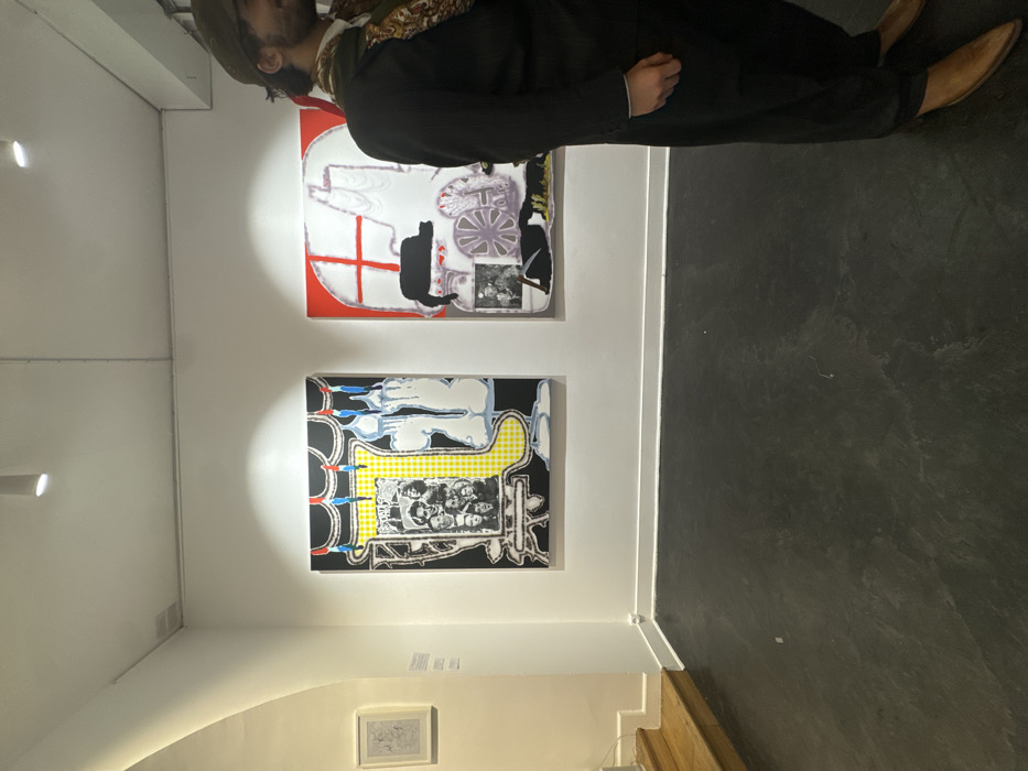 A man wearing a hat and dark clothing views artwork displayed on a white wall. Two large paintings dominate the frame: the upper one with a white skull motif on a red background, and the lower one featuring a black and yellow checkered pattern above a black and white portrait. A small framed drawing sits on the wall beneath the paintings. The floor transitions from a polished gray to wooden steps.