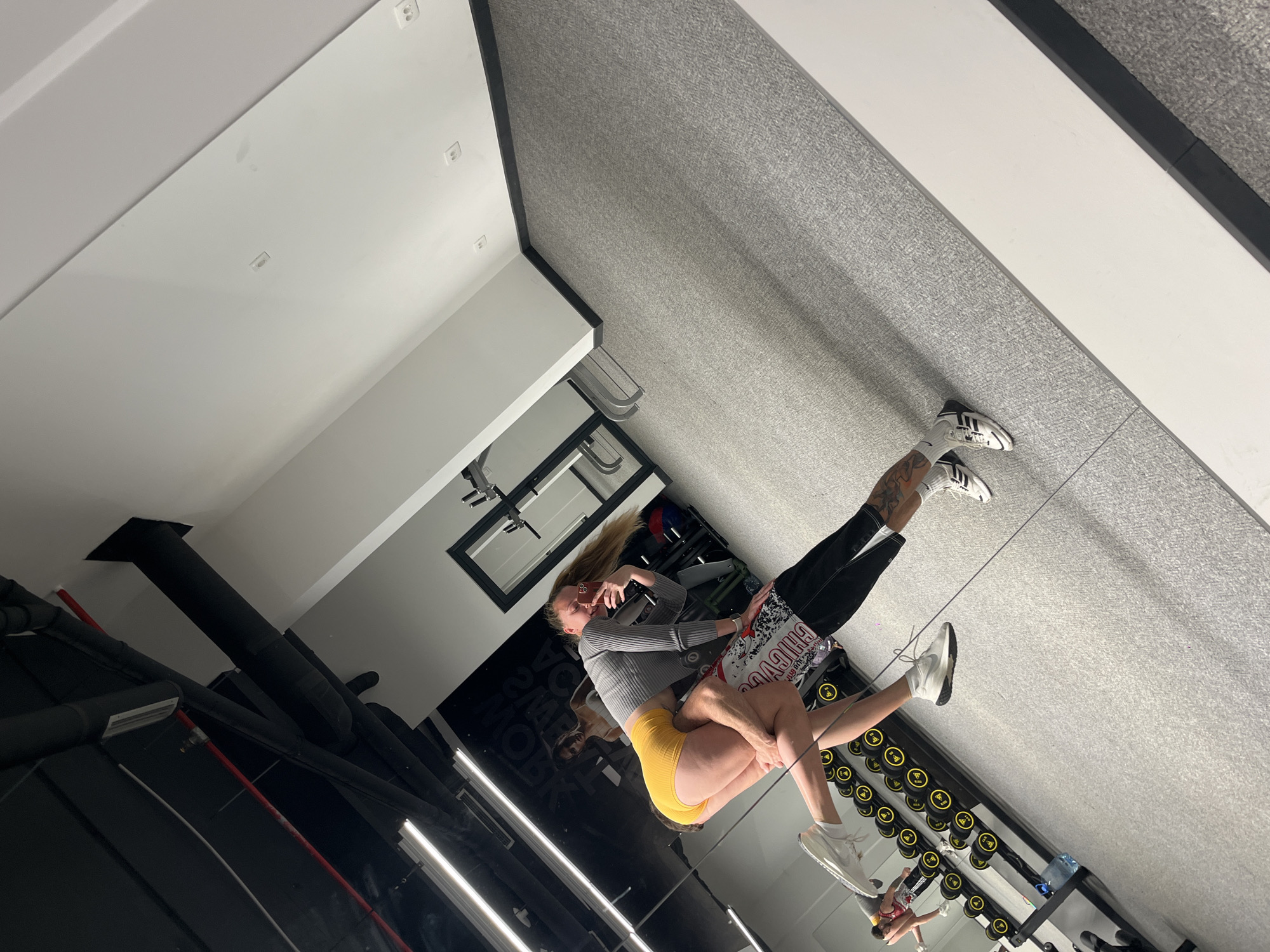 The image displays two people interacting in what appears to be a modern gym or fitness studio in Boreti, Montenegro. One individual, wearing a grey long-sleeve top and bright yellow shorts, is being lifted or carried in an embrace by another person. The second individual wears black pants or leggings, white socks, and black and white Adidas sneakers. Partially visible under the first person are graphic shorts on the second individual, featuring the text "CHICAGA" (likely referring to "CHICAGO").
The setting features a prominent mirrored wall on the right, which reflects the subjects and a long rack filled with black and yellow dumbbells. The floor is a light grey, textured material, possibly carpet or gym flooring, with a clean white strip running along the base of the mirrored wall. The ceiling is white with multiple recessed square lights providing bright illumination.
In the background, visible through the reflection and directly on the left, are black structural elements and additional gym equipment, including what looks like a pull-up bar frame. A cropped detail reveals exposed black and red pipes or ventilation ducts near the ceiling, suggesting an industrial design aesthetic. The reflections create a dynamic and multi-layered view of the scene, with blurred text appearing on the mirrored surface, partially decipherable as "POWER ZONE GYM". The overall atmosphere suggests a moment of playful interaction or partner fitness activity within a well-lit, contemporary gym environment.