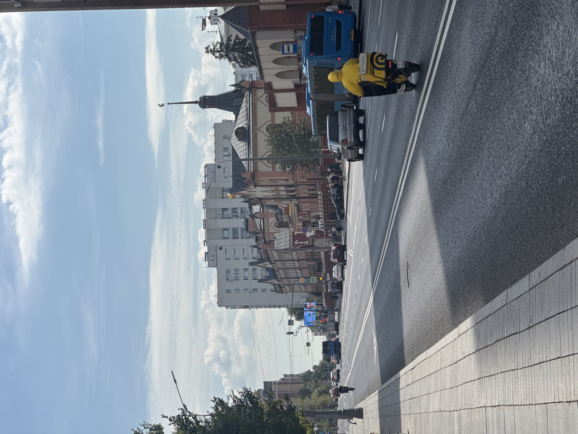 The image captures a street scene in Moscow, Russia, on a sunny day with scattered clouds. The perspective is from the sidewalk, looking down a wide asphalt road with double white lines indicating lanes of traffic. On the right side of the road, a yellow delivery scooter with a large yellow insulated delivery box on the back is moving away from the camera. The scooter has a license plate that reads "УХ1 68Х". The rider is wearing a yellow hooded jacket. To the right of the scooter, a blue bus is also traveling in the same direction. Further down the road, there are several cars, including a silver SUV.
The left side of the street features a gray tiled sidewalk, shaded by a tall tree. Beyond the sidewalk, there are buildings. The buildings closest to the road on the right are in a distinct architectural style with pink and red facades, ornate decorations, and dark gabled roofs. One building has a tall, dark spire topped with a weathervane. Further back, larger, modern apartment buildings with white facades and numerous windows are visible. A large digital billboard displaying an advertisement with a smiling man is positioned over the road. Traffic lights and street lamps are also present. People are seen walking on the sidewalk in the distance, and a cyclist is riding on the road. The overall impression is of a busy urban environment.