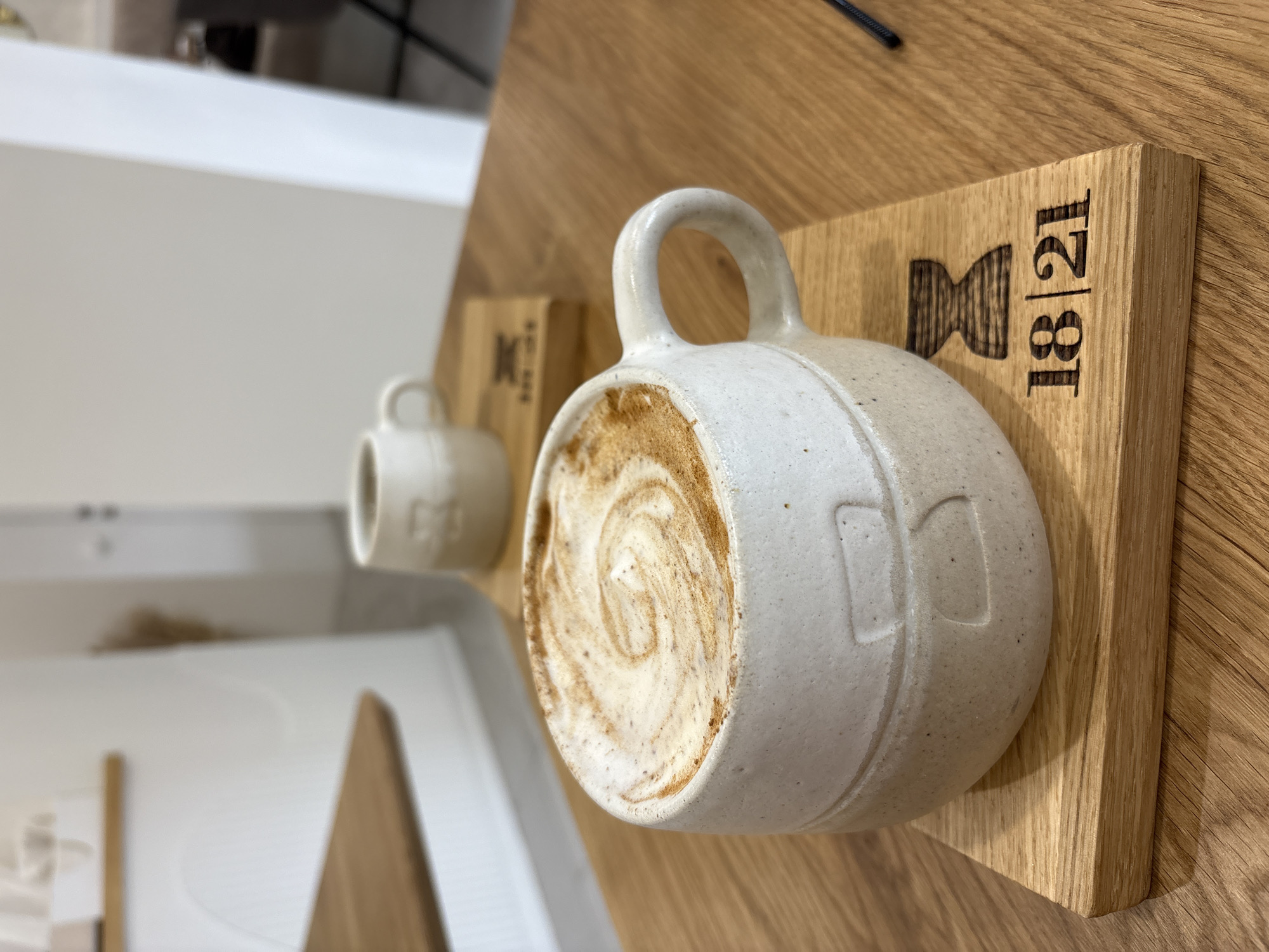 The image shows two ceramic mugs on wooden boards, presumably in a café or restaurant. The larger mug is filled with a cappuccino, latte or similar coffee drink, topped with latte art. The smaller mug sits slightly out of focus behind it. The wooden boards are branded with a logo, including the number "18|21". The overall setting seems to be modern and minimalist, with a white and wooden color scheme. The specific location, Krasnoyarsk, Russia, cannot be confirmed based solely on the image content.