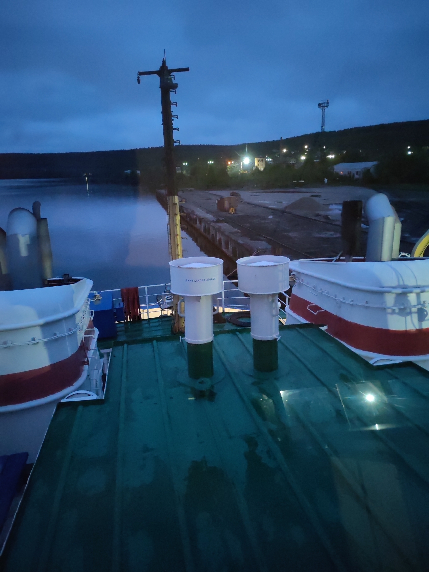 The image captures a twilight or early night scene from the deck of a vessel moored at a dock in Medvezhyegorsk, Russia. The deck, appearing wet and reflecting light, features two white and red lifeboats or small tenders, and two tall, white cylindrical ventilation structures. One of these structures prominently displays the Cyrillic text "АККУМУЛЯТОРНАЯ," which translates to "BATTERY ROOM" or "ACCUMULATOR." A mast-like structure rises from the vessel on the left.
Beyond the vessel, a calm body of water, possibly a lake or bay, stretches towards a wooden pier. The water's surface is dark and reflective, with a light mist or fog hovering above it. Along the distant shore, a settlement is visible, nestled against a dark, forested hill. Numerous artificial lights from buildings and streetlights dot the landscape, creating warm glows against the cool blue, overcast sky. A tall, industrial-looking tower stands on the hillside, overseeing the settlement.
The time of day is clearly evening or early night, characterized by the deep blue sky and reliance on artificial illumination, suggesting either dusk, early morning, or the prolonged twilight of northern latitudes. No people are visible, contributing to the quiet and serene atmosphere. The overall impression is one of peaceful stillness at a northern harbor.