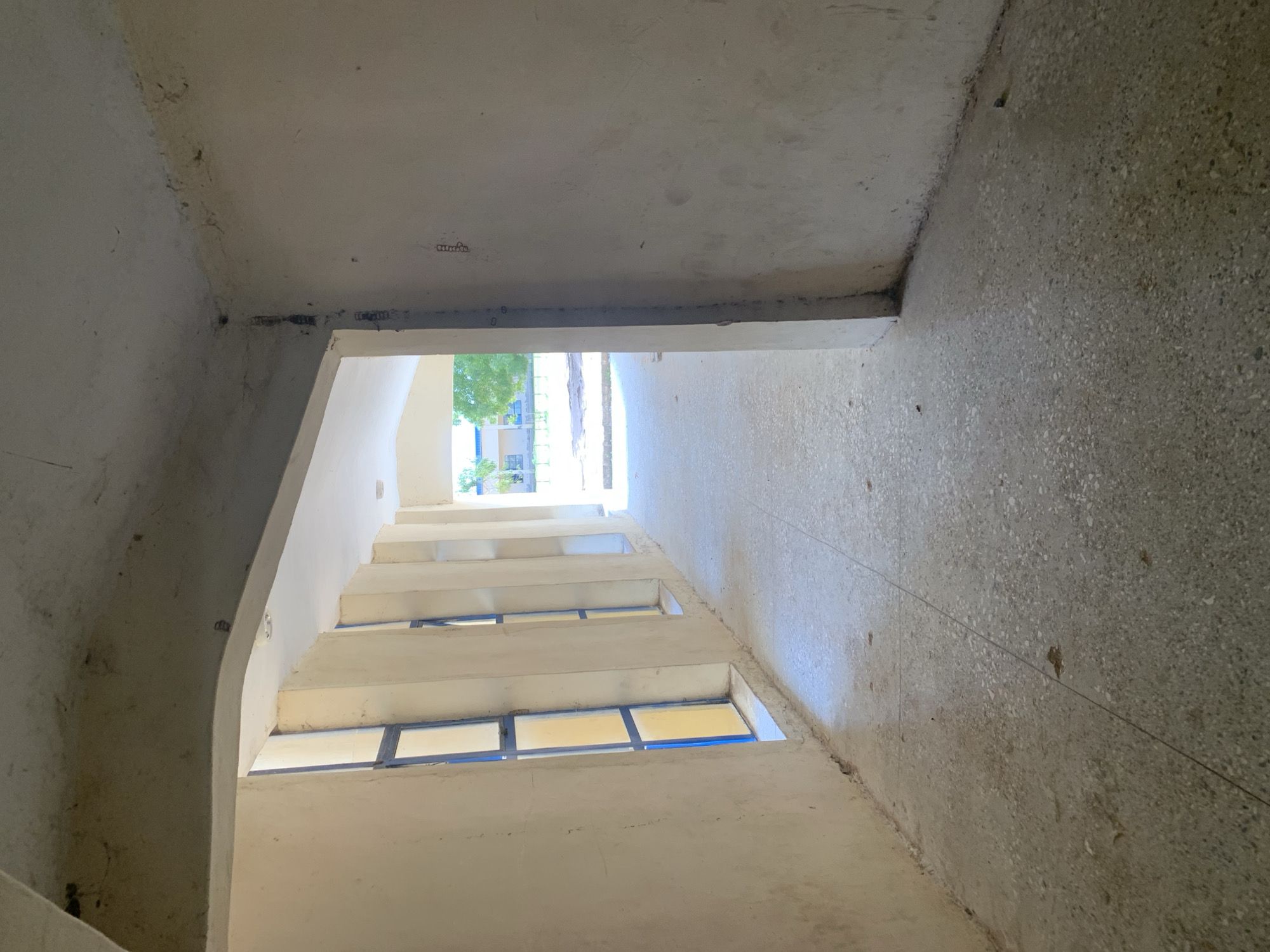 The image depicts an outdoor corridor with a polished concrete floor and pale yellow walls. Tall, narrow windows with blue frames and yellow glass are set into the left wall. The corridor extends into the distance, where trees and a building are visible under a bright sky. The overall impression is one of an unfinished or weathered architectural space. The scene is located in Jalingo, Nigeria.