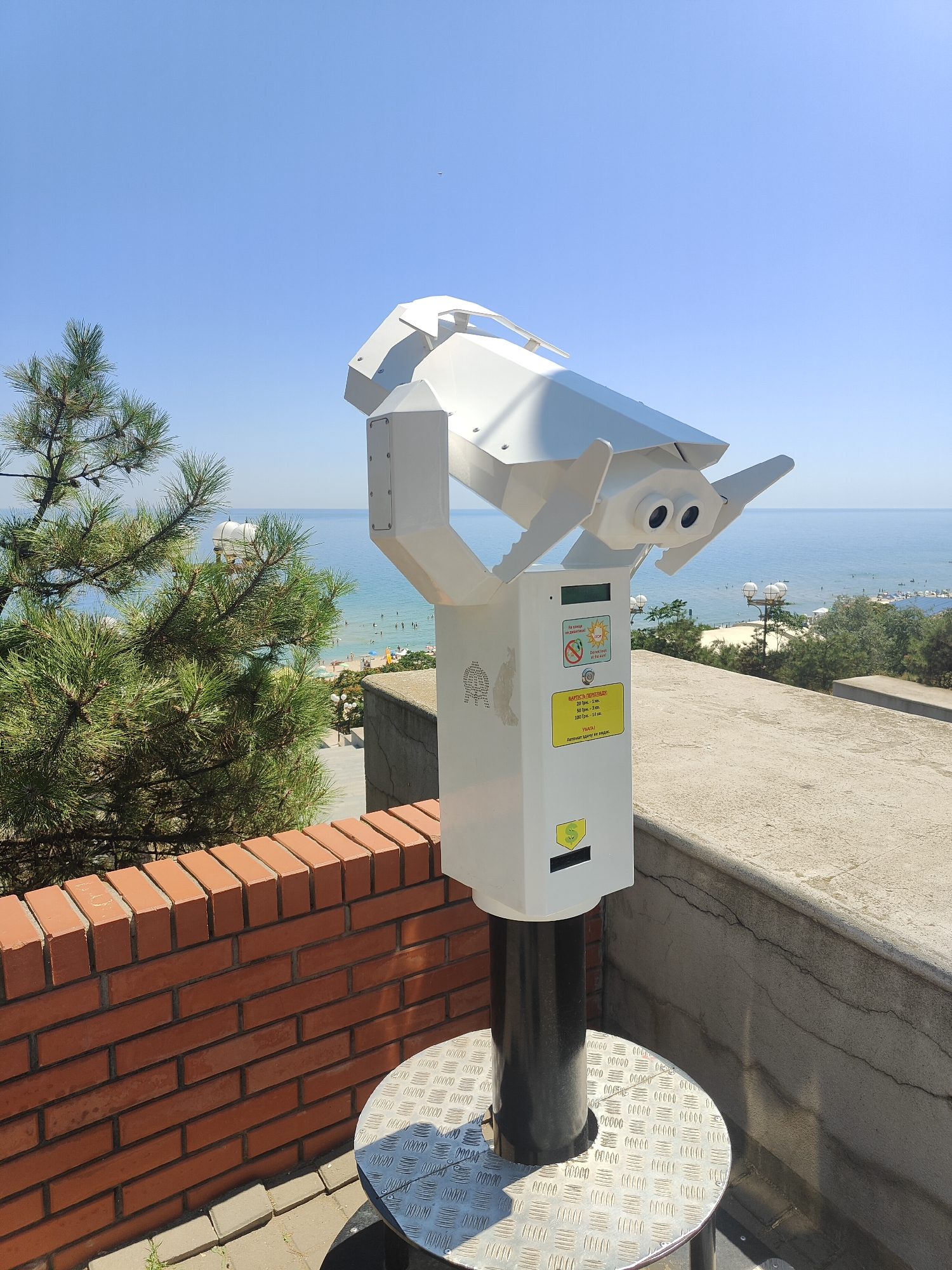 The image shows a view of the Black Sea from an elevated position in Chornomorsk, Ukraine. In the foreground, a white, modern-looking coin-operated binocular viewer stands on a metal base. The viewer is pointed towards the sea. Behind the viewer, a red brick wall is visible, topped by a concrete railing. To the left of the viewer, a pine tree with cones is in the frame. In the mid-ground, the Black Sea stretches out, with a beach lined with people, umbrellas, and buildings. The sky is clear and blue. The overall scene suggests a sunny day at a seaside resort.