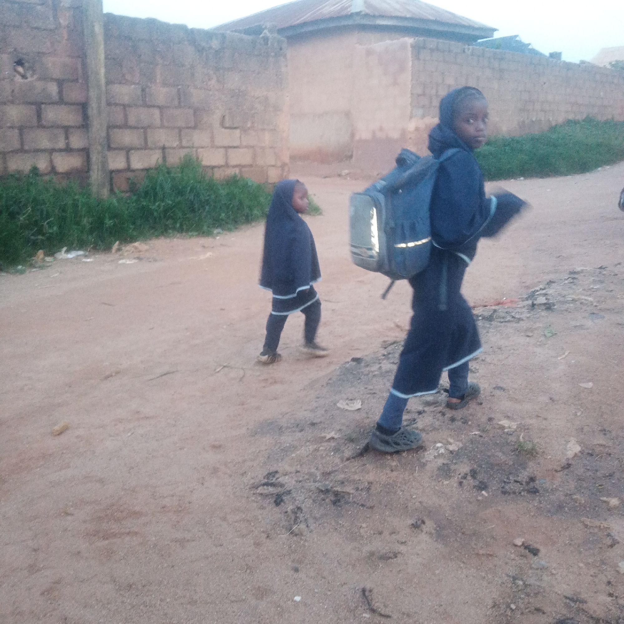 Two young girls, presumably students, walk along a dusty path in Jos, Nigeria. The girl on the right, facing away from the camera, wears a dark blue headscarf and a long blue garment with a light blue trim. She carries a dark blue backpack with reflective strips. The girl on the left, also facing away, wears a similar dark blue headscarf and a long dark blue garment with a white and light blue trim over dark leggings. She walks ahead of the other girl, her back to the camera. The environment is characterized by a dirt road with scattered debris, a weathered brick wall on the left, and a simple building with a corrugated metal roof in the background. Lush green vegetation grows along the base of the wall. The lighting suggests it is either early morning or late evening.