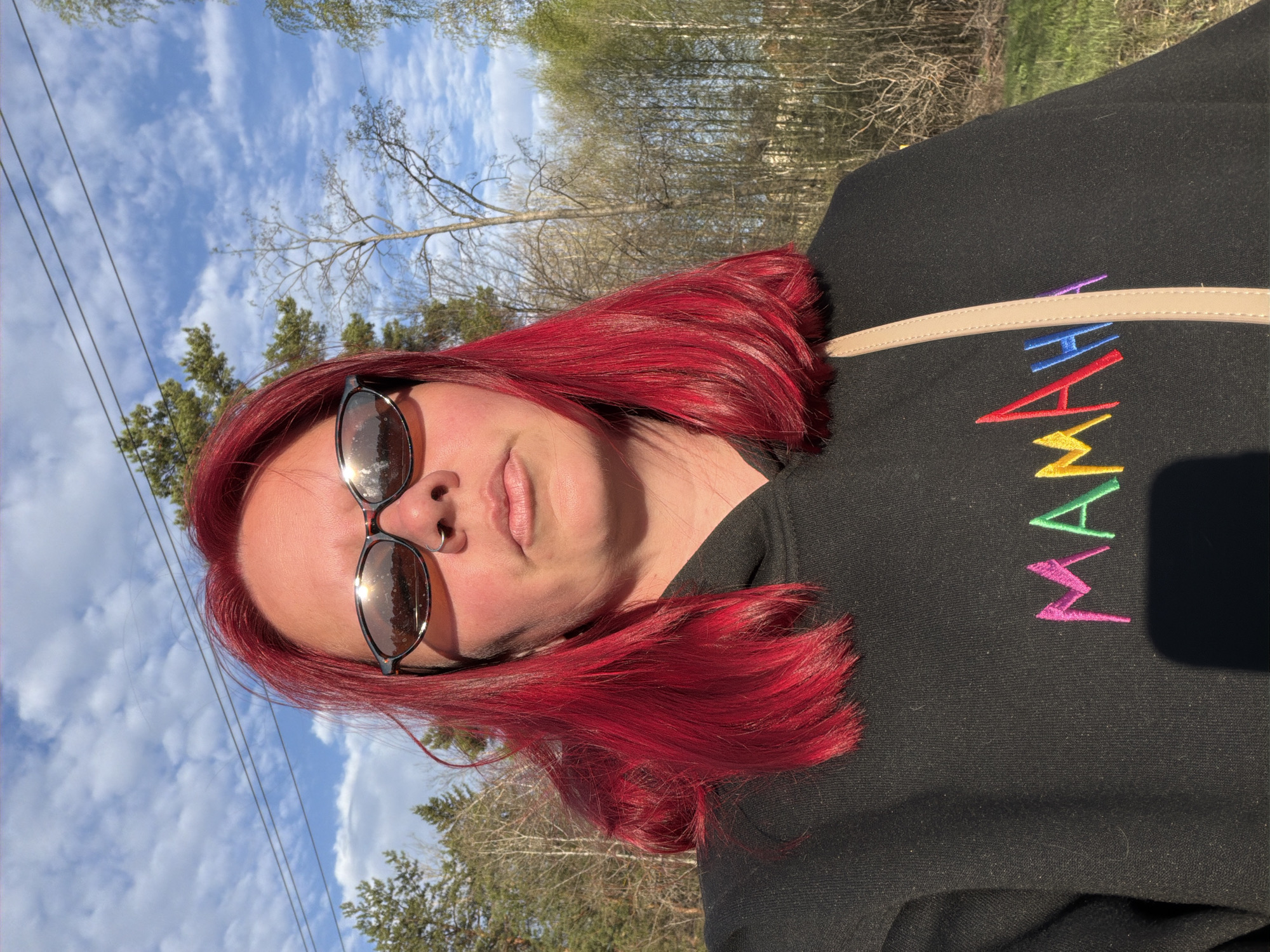 A woman with red hair and sunglasses is pictured outdoors. She is wearing a black sweatshirt with the word "MAMA" embroidered in rainbow colors. She has a nose ring on her left nostril. The background shows trees, a partly cloudy blue sky, and telephone wires. The location is inferred to be in the city of Chaikovsky, Russia based on the prompt.