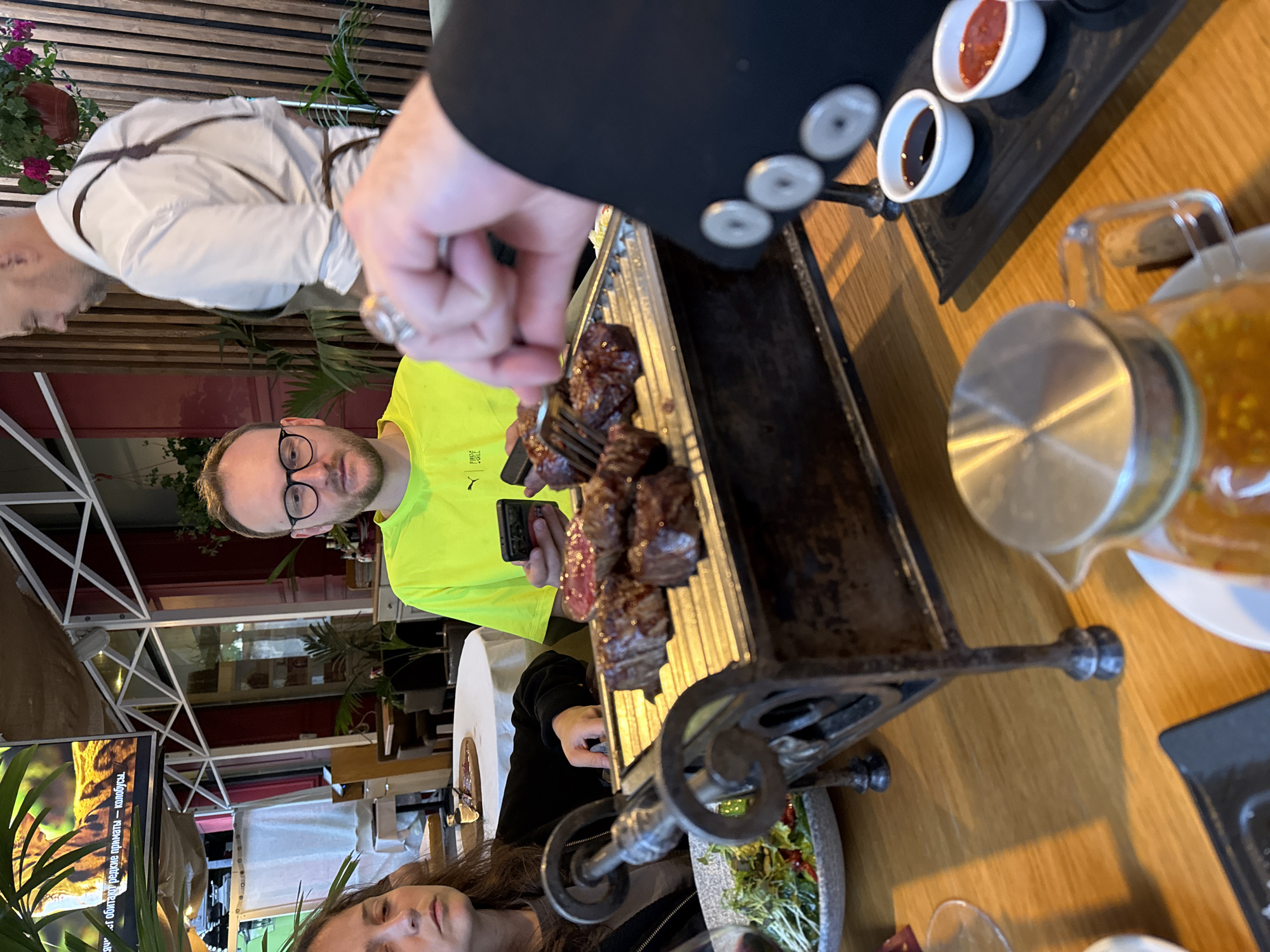 Scene: Interior of a restaurant setting, possibly in Moscow, Russia.
People: Three individuals are partially visible. One man, seen from the chest up, wears a bright yellow t-shirt and glasses, holding a cell phone. A woman with long brown hair is visible in the bottom of the frame. Another person wearing a white jacket can be seen on the top left of the frame.
Objects: A serving of grilled meat sits on a metal grill with decorative wrought iron elements. Accompanying the meat are small dishes containing sauces, and a glass pitcher filled with liquid, possibly juice. A plate with a salad is also present.
Actions: A person in a black suit is using a fork to pick pieces of meat off the grill. The man in the yellow shirt appears to be taking pictures or videos with his cell phone.
Environment: The restaurant appears to have an indoor/outdoor feel with white metal framing visible and glimpses of greenery through the windows, implying a location with potentially high tourism activity.