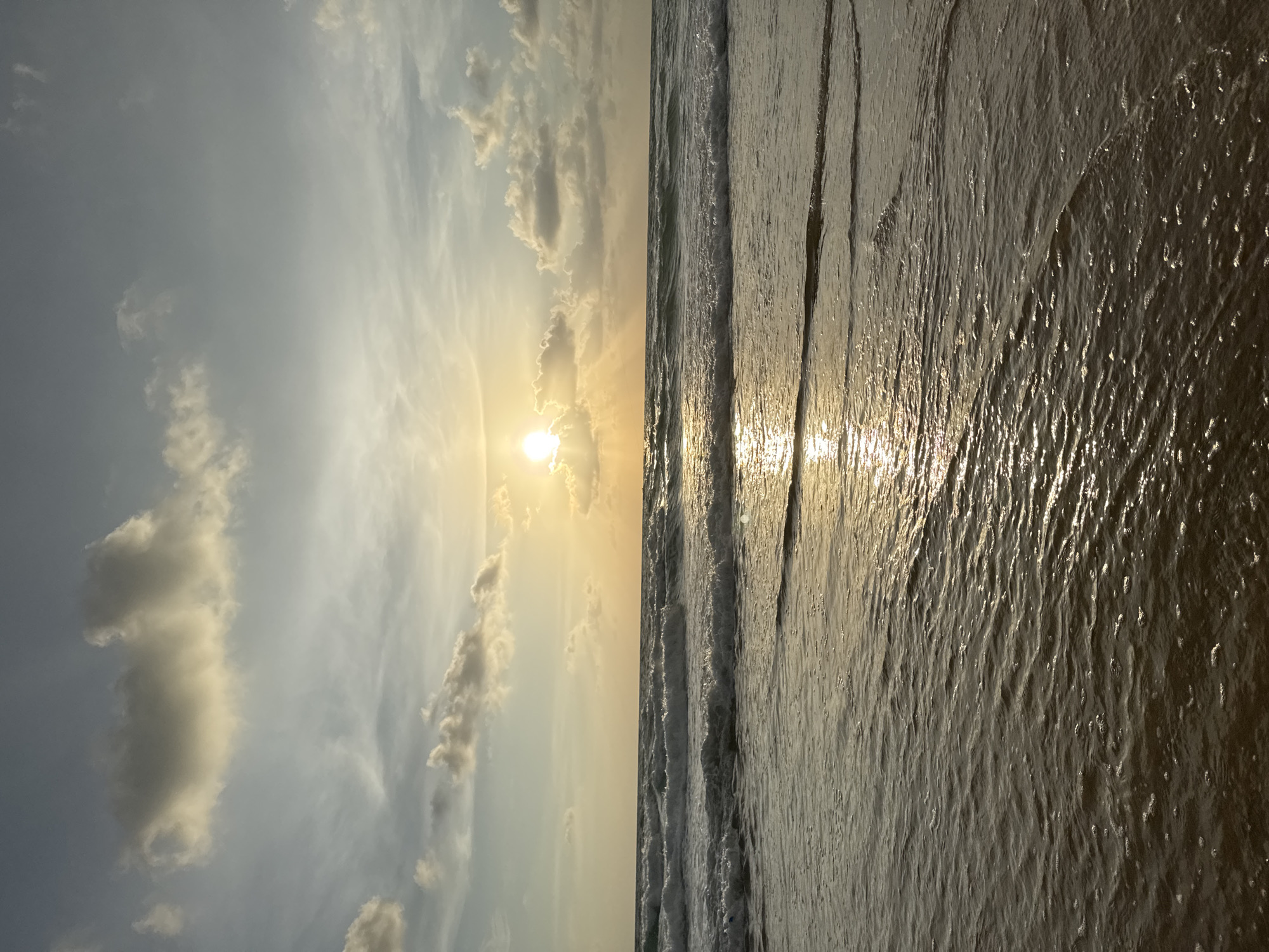 An outdoor, medium shot captures a tranquil beach scene at sunset in Karon, Thailand. The sun, a bright, white orb partially obscured by clouds, is positioned in the upper center of the frame, casting a warm, golden glow across the sky and water. Rays of sunlight emanate from behind the sun, creating a radiant halo effect. The sky is a gradient of soft blues and yellows, with scattered, wispy clouds tinged with the warm hues of the setting sun. Below the sky, the ocean stretches out, with gentle waves rolling towards the shore. The water is a deep blue-green, reflecting the sunlight in shimmering patterns. Foamy white crests adorn the tops of the waves, indicating their movement. The foreground is dominated by the wet sand of the beach, glistening with a thin layer of receding water. Small ripples and bubbles are visible on the surface of the water near the shore. No people or animals are present in the image. The overall atmosphere is one of peace and serenity.