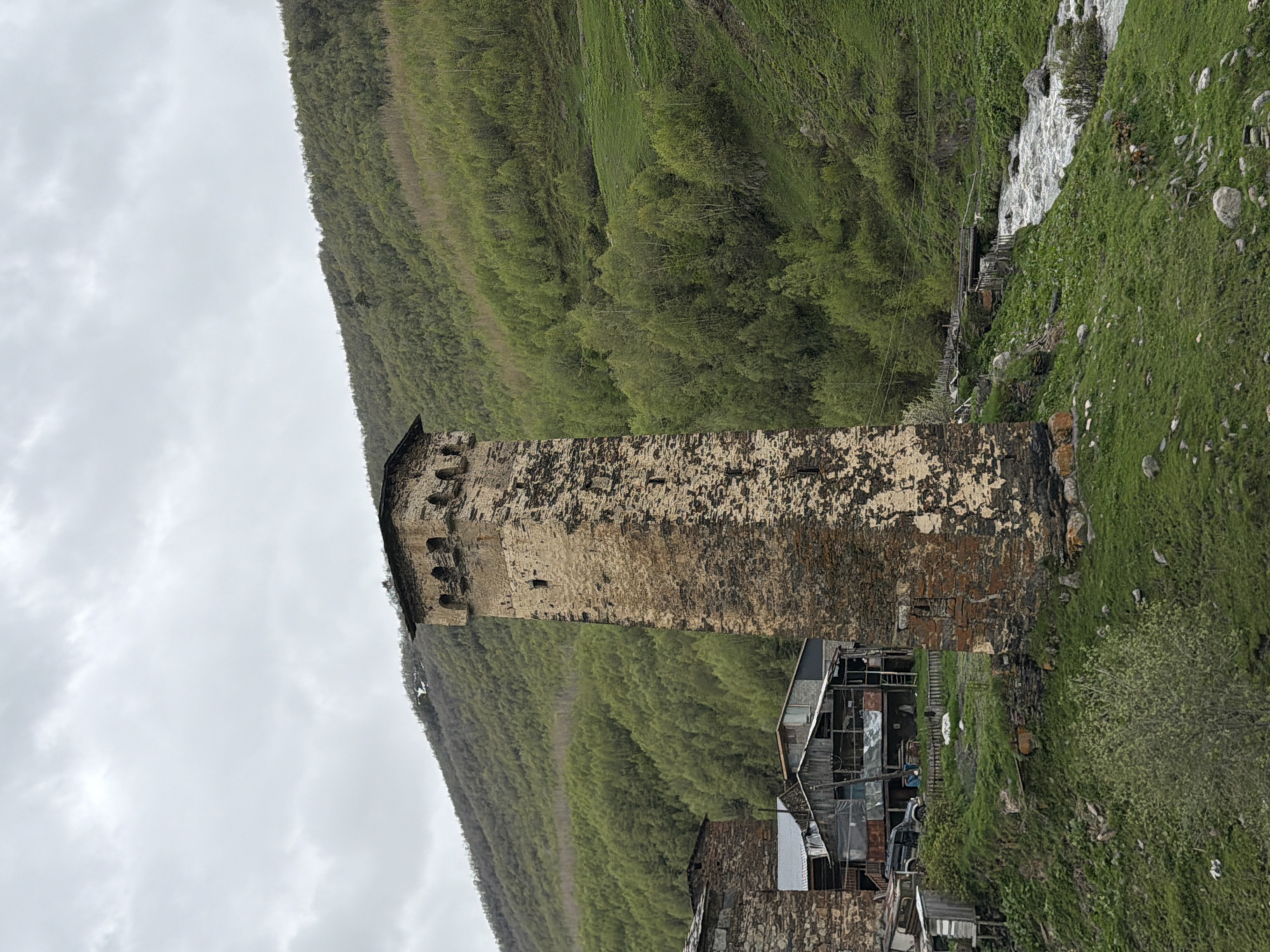 The image shows a medieval stone watchtower in the village of Ushguli, Georgia. The tower is tall and narrow, with a square base and a flat roof. It is made of rough-hewn stones and has small windows at various levels. The tower is surrounded by other stone buildings, some of which appear to be residential houses. A river flows in the foreground, and a lush green hillside covered in trees rises behind the village. The sky is overcast, suggesting a cloudy day. There are no people visible in the image. The scene depicts a remote and historic settlement nestled within a mountainous landscape.