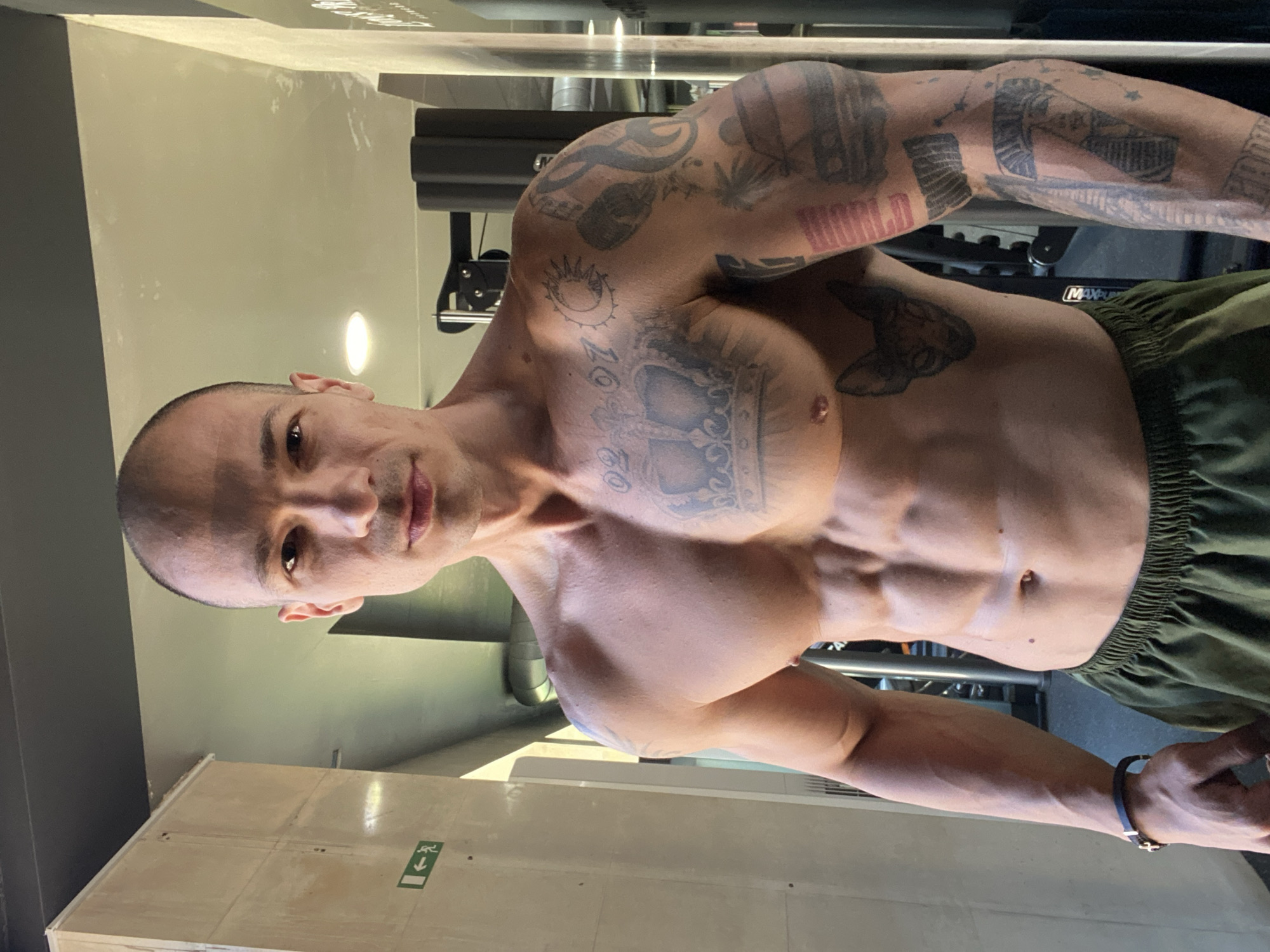 A shirtless man with extensive tattoos on his torso and arms stands facing forward in a gym. He has a shaved head and is wearing green athletic shorts. The background shows gym equipment and a textured wall. The image is a medium shot, focusing on the man's upper body. There is no identifiable information or context linking this image to the city of Pleven, Bulgaria.