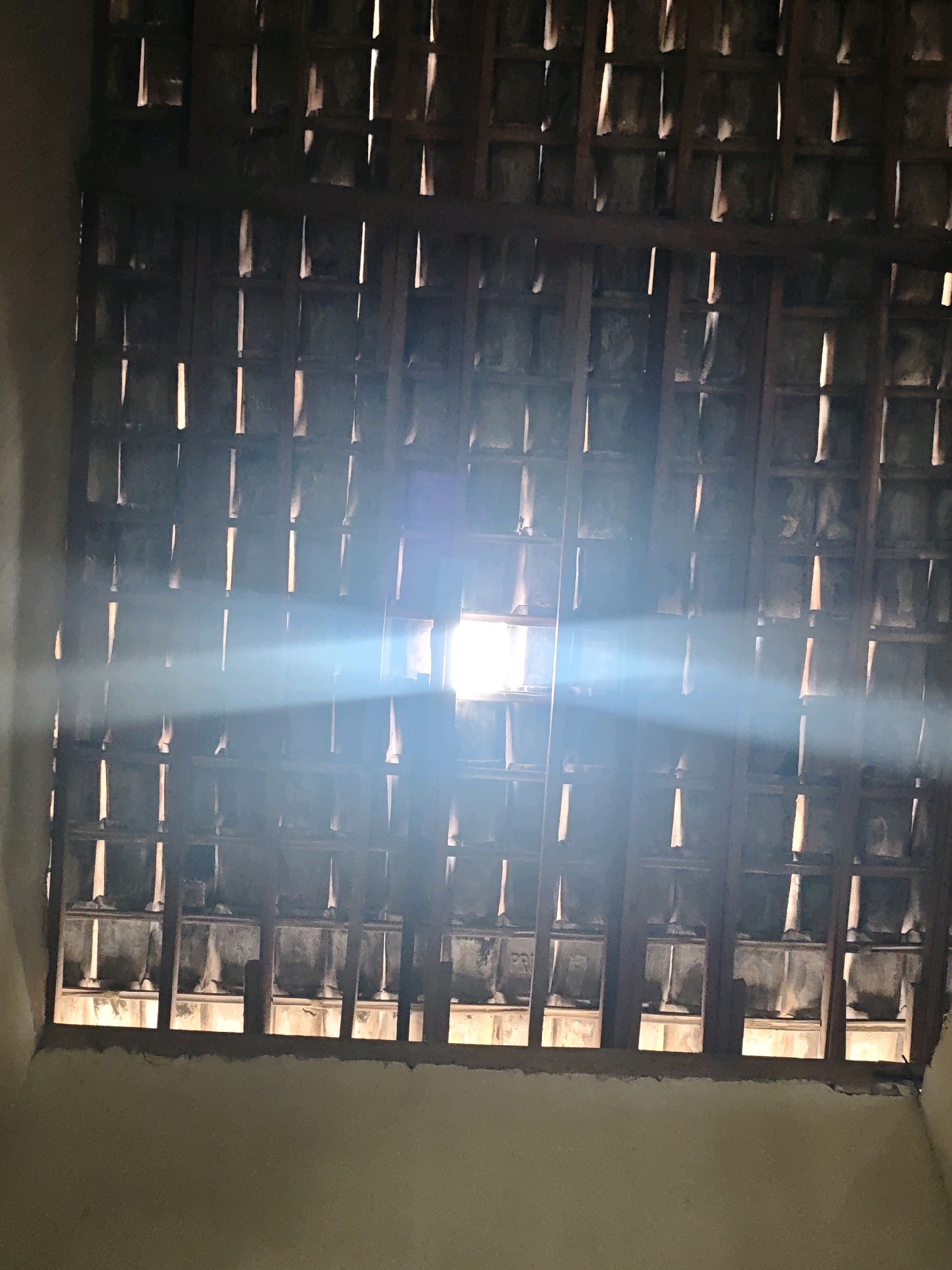 The image shows a close-up view of a window with a wooden lattice structure. Sunlight streams through the gaps in the lattice, creating a bright, diffused light that illuminates the interior. The lattice is composed of dark wooden beams arranged in a grid pattern, with individual square or rectangular openings. Through these openings, a textured, tiled surface is visible, suggesting a roof or ceiling structure. The overall impression is one of a dimly lit space, possibly a room or building in Pagu, Indonesia, where natural light is filtered through a traditional architectural element. The light beam is a prominent feature, cutting across the frame and highlighting the dust or particles in the air. The surrounding walls are a muted, light-colored material. No people or specific objects are identifiable within the scene, other than the architectural elements of the window and the light itself.