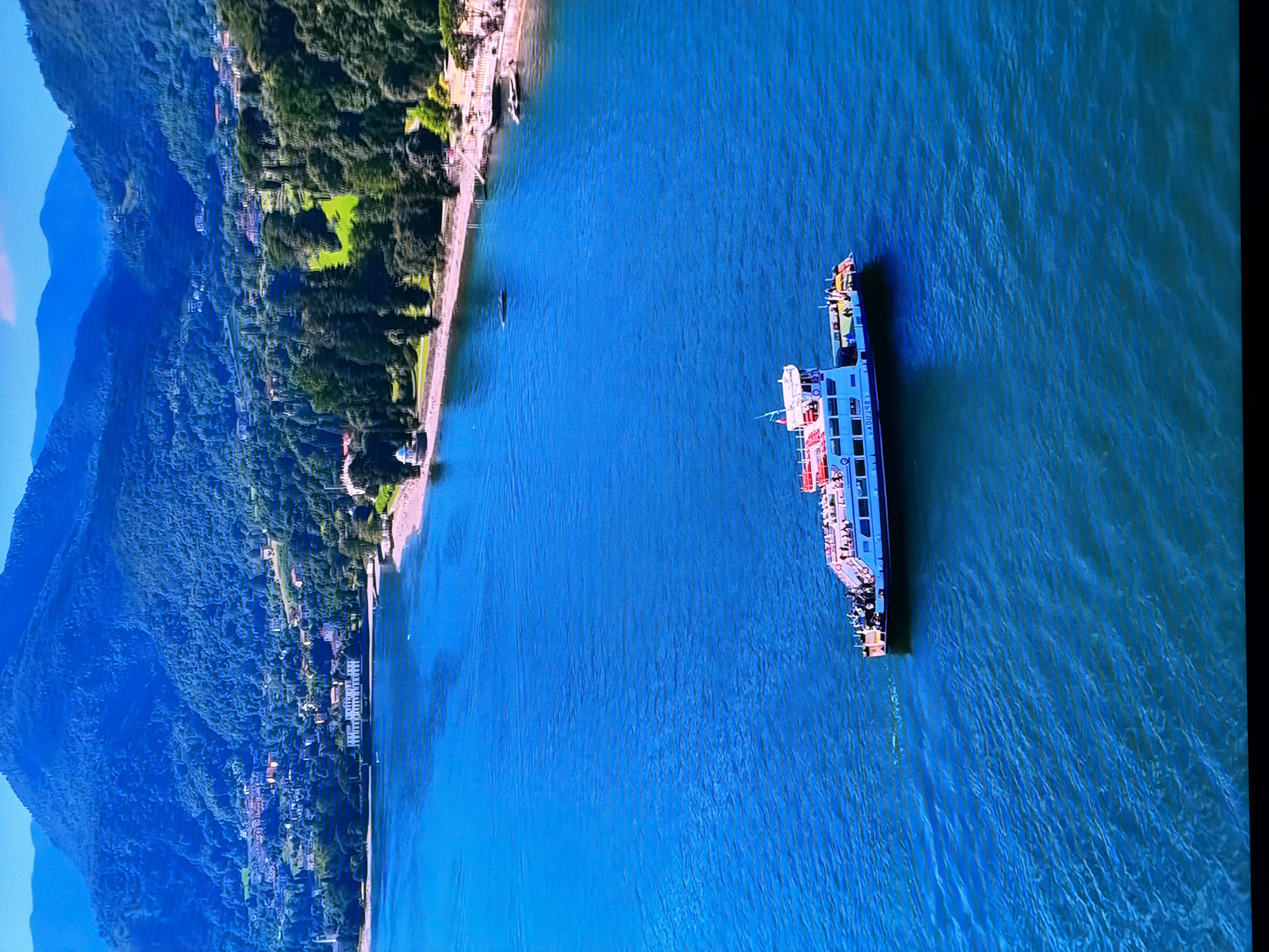 The image is an aerial view of a wide, deep blue lake with a ferryboat in the foreground. The ferryboat is blue and white with red accents and appears to be carrying passengers on its upper deck. The water is choppy with visible ripples. In the background, there is a shoreline with trees, buildings, and mountains. The mountains are covered in dense green foliage. The buildings along the shore appear to be a mix of residential and possibly public structures. The scene depicts a sunny day with clear skies. There is no visible connection to Chirchik, Uzbekistan.