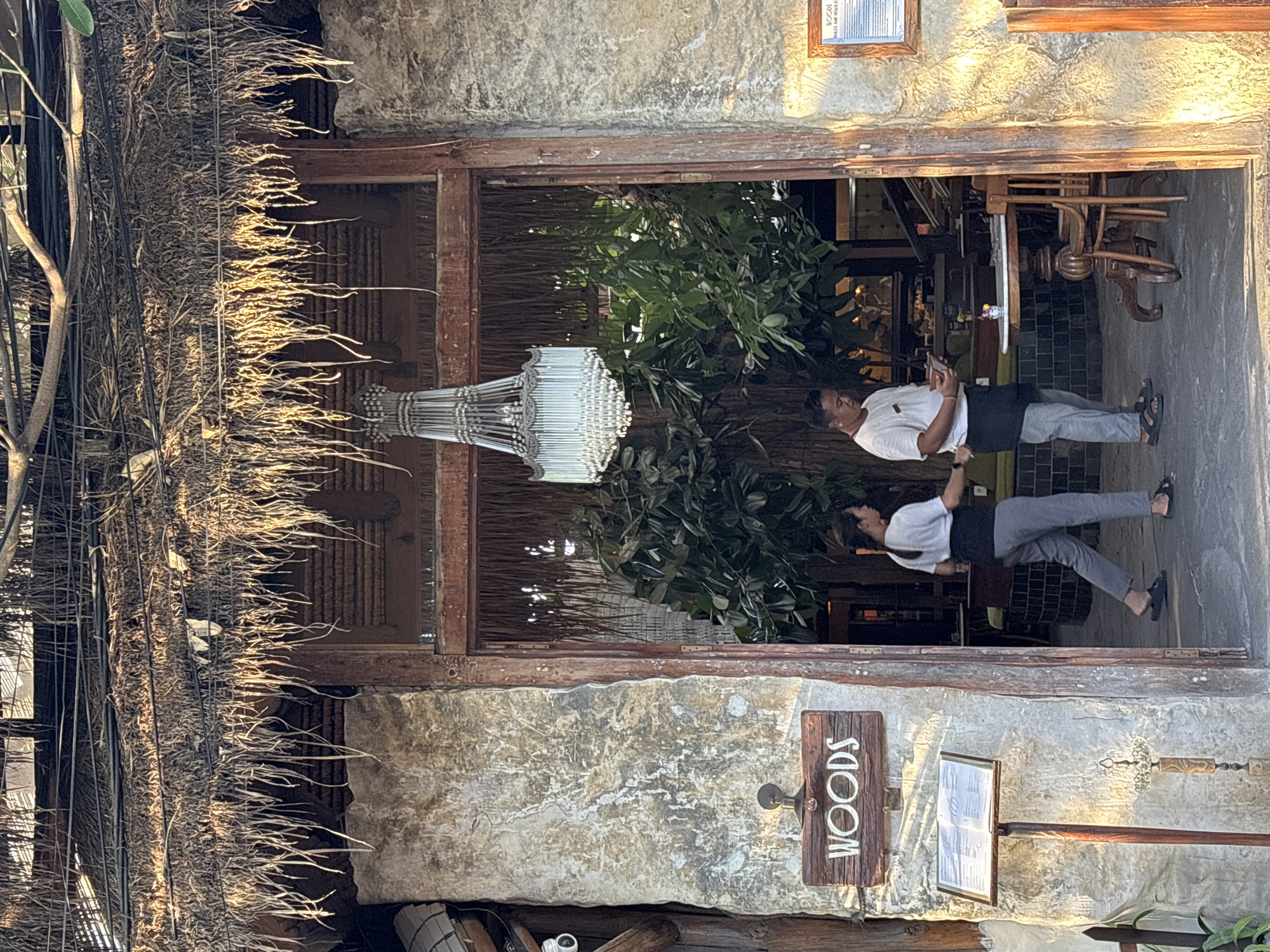 The image captures the entrance of the "Woods" restaurant in Mengwi, Indonesia. The entrance is framed by a rough, stone-like facade and a rustic thatched roof. A sign bearing the name "Woods" is affixed to the facade. A menu stand sits beside it. Inside the entrance, two waiters are visible, dressed in white shirts and dark pants or skirts. They are standing near a counter, seemingly interacting with customers or preparing for service. A decorative chandelier hangs inside the entrance. The interior features greenery and furniture, hinting at the restaurant's ambiance.