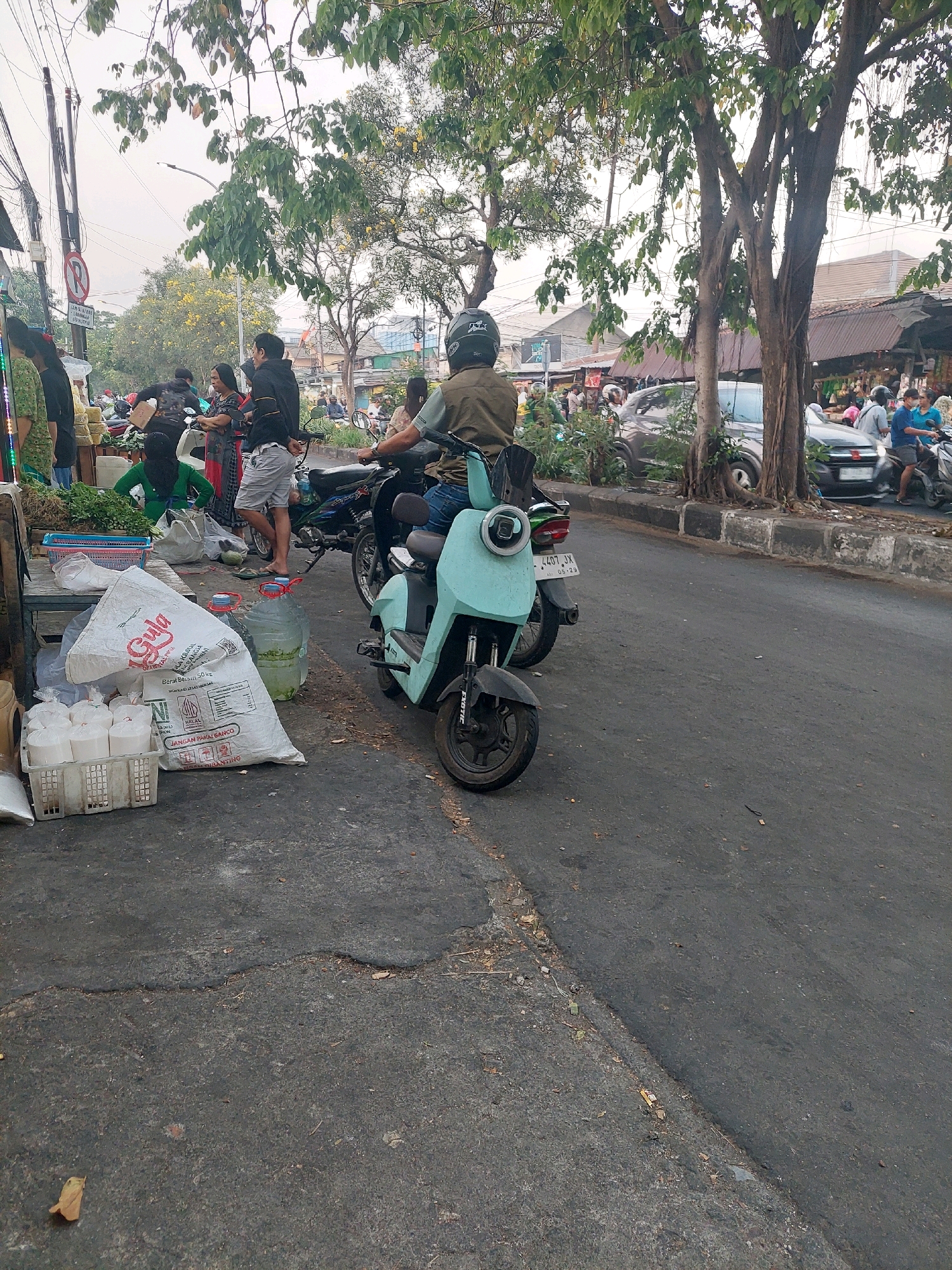 In Surabaya, Indonesia, on a cloudy day, a street scene unfolds with a focus on a light blue electric scooter. The scooter, with license plate "4407 JX" and a date "05.29" visible, is parked on the asphalt road. A person wearing a dark helmet and a khaki vest rides the scooter, facing away from the camera. To the left of the scooter, a large white bag of sugar with Indonesian text is placed on the ground, alongside several plastic containers filled with a clear liquid. Smaller white plastic containers are stacked in a white crate next to the sugar bag.
The background reveals a busy street with other motorcycles and cars. Various vendors are visible on the left side of the road, displaying produce and other goods. People are seen walking and interacting, suggesting a market or shopping area. Trees with dense foliage line the street, casting shadows. Buildings with corrugated metal roofs are present in the distance. The overall environment appears to be an outdoor market or a bustling urban street with commercial activity.