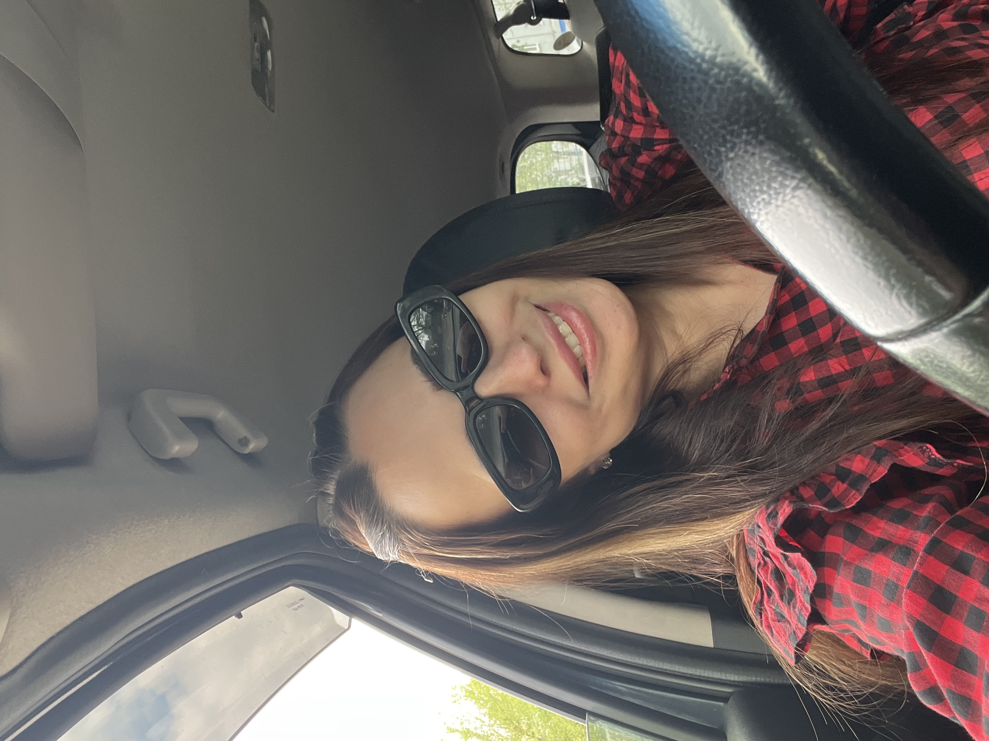 A woman with long brown hair and sunglasses is inside a car. She is wearing a red and black checkered shirt and smiling. The car's steering wheel is visible, as well as the interior ceiling and a window showing greenery outside. The location is Unknown, Unknown.
