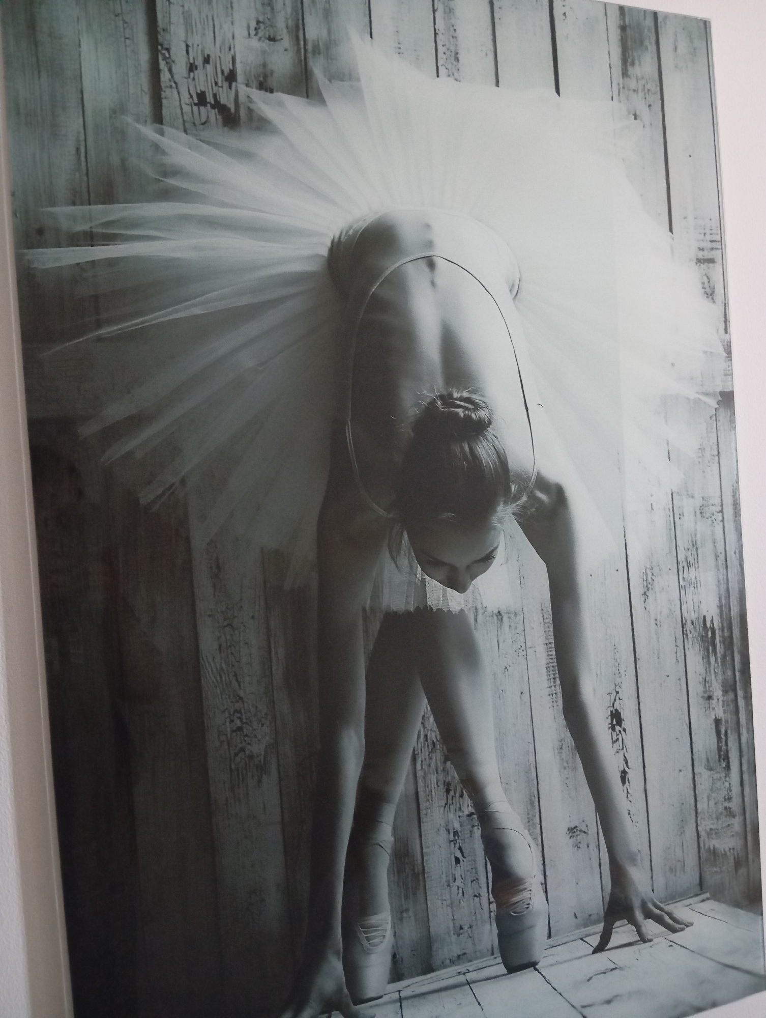 The image is a black and white photograph of a ballet dancer in a tutu, performing a graceful pose against a backdrop of weathered wooden planks. The dancer is bent over, with her arms extended and her head lowered, showcasing the flexibility and elegance of her form. Her hair is neatly tied in a bun. The lighting in the image is soft, highlighting the textures of the tutu and the wood. The overall mood is serene and artistic. The image is framed with a white border and appears to be a piece of art mounted on a wall. There is no discernible information about its location in Dubai, United Arab Emirates.