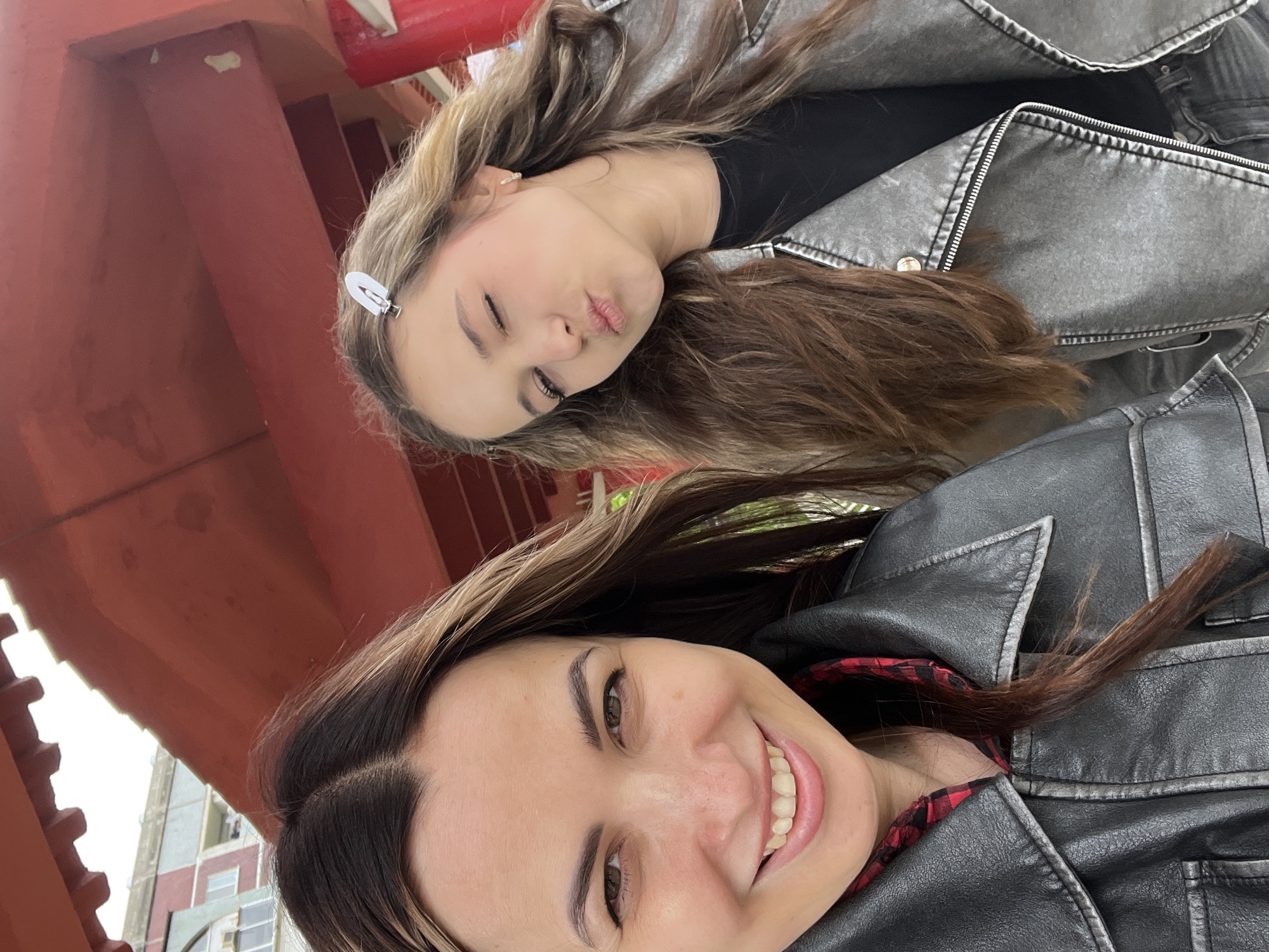 Two women are posing for a selfie. The woman in the foreground is smiling and wearing a black leather jacket with a red plaid shirt underneath. The woman in the background is making a kissing face and wearing a grey leather jacket and a black shirt. She has a white clip in her hair. They are both standing under a red structure, possibly a bridge or building overhang. Buildings are visible in the background. The city is unknown.