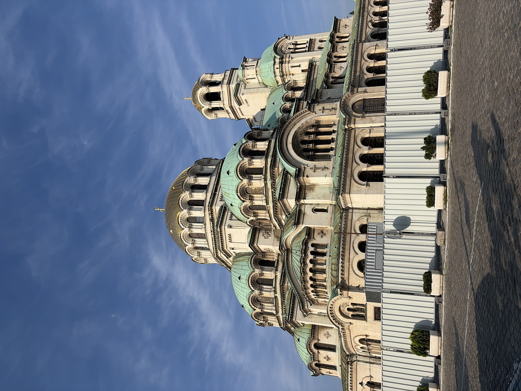 The image displays the Alexander Nevsky Cathedral in Sofia, Bulgaria, a large Neo-Byzantine structure. The cathedral occupies the majority of the frame, showcasing its multiple domes and intricate facade under a clear blue sky with sparse, wispy white clouds.
The central and largest domes are golden/bronze in color, topped with golden Orthodox crosses. Numerous smaller domes and roofs are covered in a distinctive verdigris (greenish-turquoise) material. The cathedral's walls are constructed from light-colored stone, featuring multiple arched windows, decorative cornices, and architectural details across several tiers. A prominent bell tower, also topped with a golden dome and cross, is visible on the right side of the main structure.
In the foreground, a temporary white corrugated metal construction barrier runs horizontally, partially obscuring the lower sections of the cathedral's base. In front of this barrier, several white rectangular planters contain small green shrubs. The ground level consists of dark, patterned paving stones, which exhibit distinct shadows cast from the left, indicating bright sunlight from a relatively low angle. A vertical pole with a small, circular sign is positioned behind the barrier on the right side of the foreground. Several small, dark specks, consistent with distant birds, are visible in the upper sky. No people are visible in the scene.