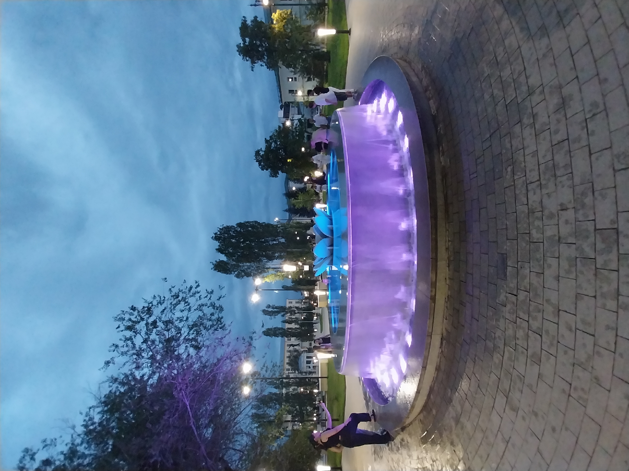 The image shows a fountain with purple and blue lights in what appears to be a park or public square. Several people are visible, some gathered near the fountain and others walking in the background. The time of day appears to be dusk or early evening, as the sky is a deep blue with some clouds, and artificial lights are on. The ground around the fountain is paved with grey interlocking stones. Trees with dark green foliage are scattered throughout the scene, and some have a purple light cast on them, possibly from the fountain. In the background, there are buildings that suggest an urban or suburban setting. One person is lying on the ground near the fountain, their body angled towards the camera, while others stand in groups. The overall mood seems relaxed and peaceful, with the illuminated fountain being a central point of interest.