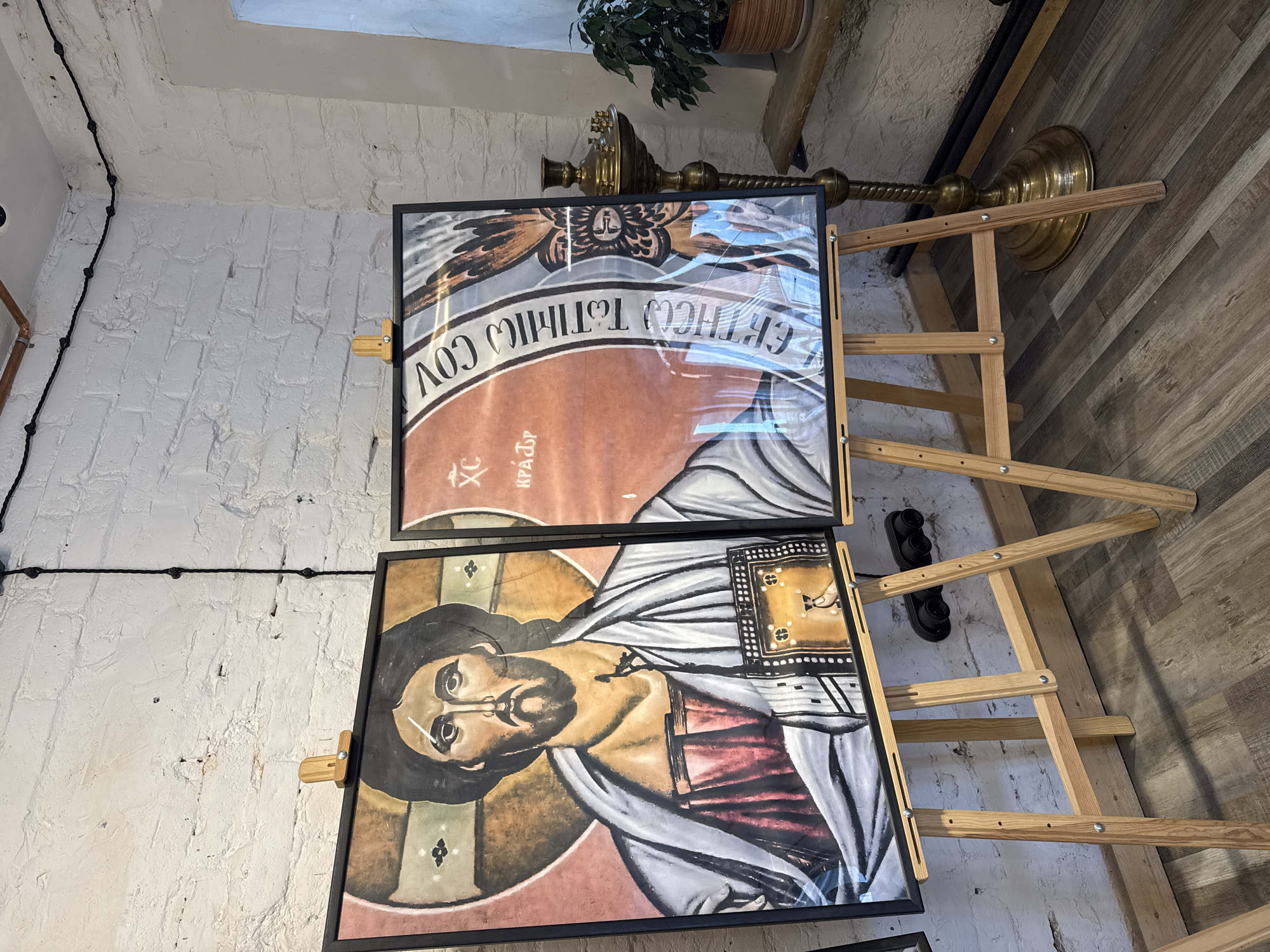 The image shows two framed icons, likely religious art, displayed on wooden easels against a white brick wall. The icons are oriented vertically and are positioned one above the other. The upper icon features a stylized depiction of a face within a circular motif, surrounded by feathered wings. There is text in a Cyrillic script visible on this icon, partially obscured by plastic sheeting. The lower icon is a portrait of Jesus Christ, with his face rendered in a traditional iconographic style, featuring dark hair and beard, and a halo. He is depicted wearing a robe and holding an open book. This icon is also covered in plastic sheeting.
In the background, to the right of the icons, is a tall, ornate brass candlestick or candelabra. Further to the right, a wooden A-frame structure, possibly part of a display or scaffolding, is visible. The floor is made of wooden planks. At the top of the image, a potted plant is visible on a shelf. The lighting suggests it is indoors, possibly a gallery, church, or workshop. The setting appears to be a rustic or minimalist interior. The overall mood conveyed by the icons is solemn and spiritual. The visible text on the upper icon reads "СВЯТИТЕЛЮ ТОЛГСКОМУ" and "ХС КРАЗОР".