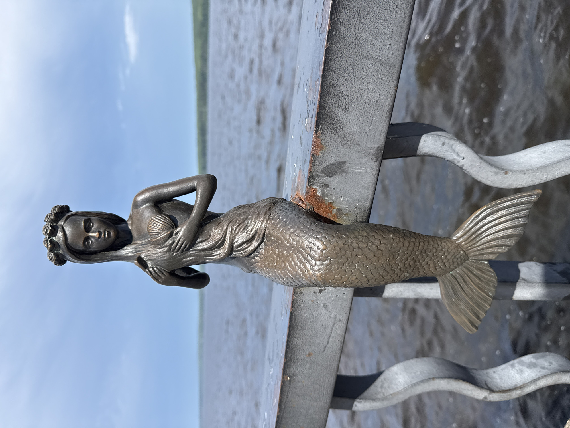 A bronze statue of a mermaid is perched on a metal railing overlooking a body of water under a cloudy sky. The mermaid is depicted with a human torso and a fish tail, holding her long hair in her hands. She wears a crown of flowers and a seashell on her chest. The railing is weathered and shows signs of rust. The water is dark and choppy, with a faint green tree line visible in the distance on the horizon. The overall scene suggests an outdoor public space near a waterfront.