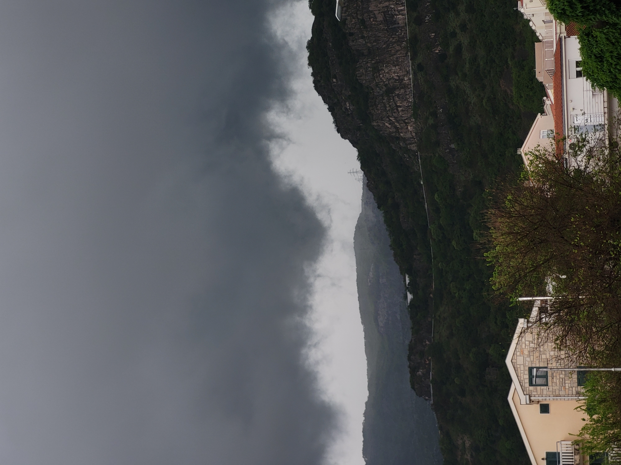 The image depicts a vertical view of a landscape scene, likely in or near Budva, Montenegro. A steep, rugged mountain range covered in dense green vegetation is visible. The left side of the image is dominated by dark, ominous storm clouds. A thin, winding road can be seen tracing along the side of the mountain. In the foreground, there are houses with beige exteriors and red-tiled roofs interspersed with green shrubbery, suggesting a residential area nestled near the mountains. The sky is overcast, contributing to a somber and atmospheric tone.