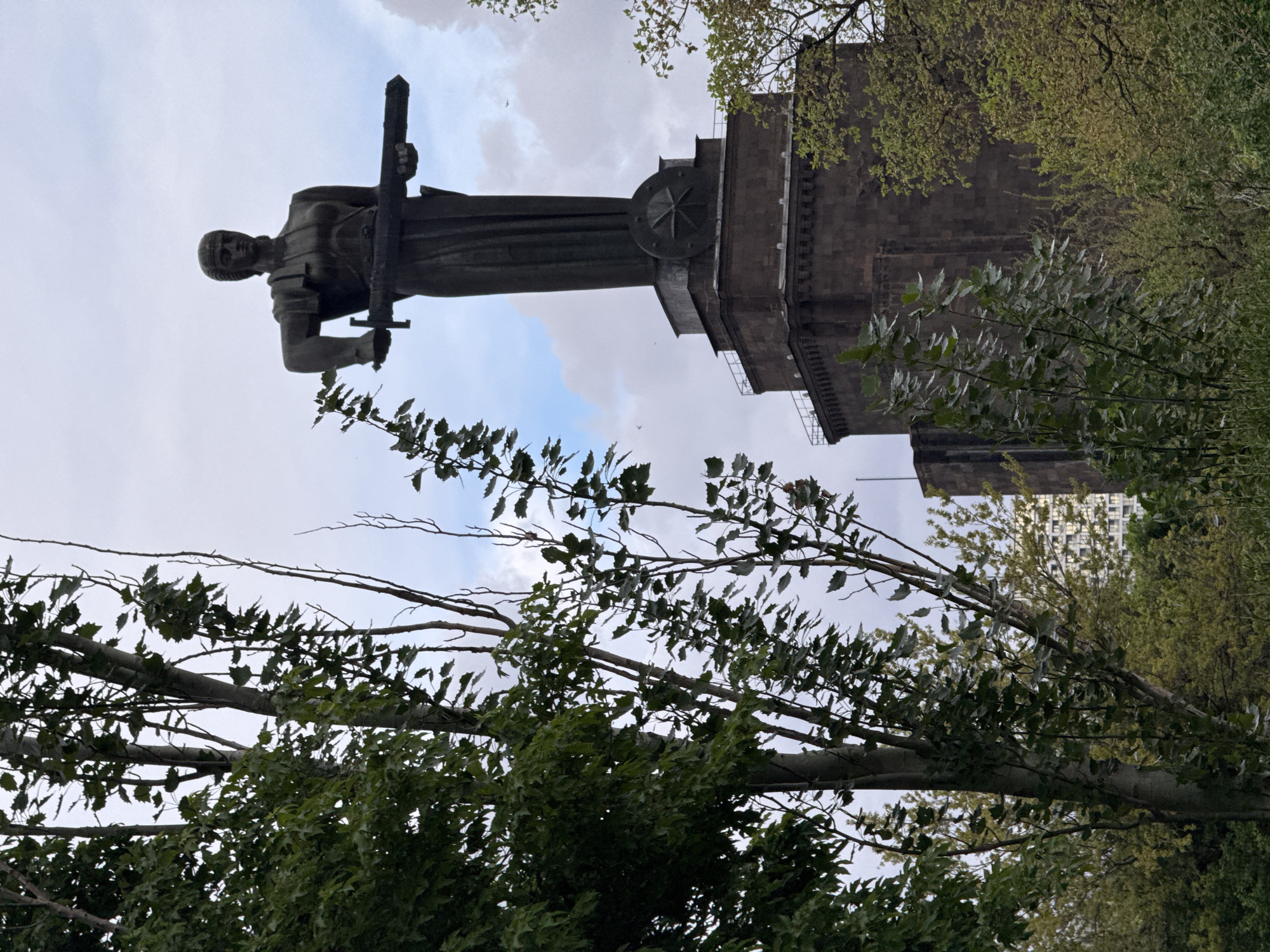 Here is a forensic description of the image:
The image shows the Mother Armenia statue in Yerevan, Armenia. The statue depicts a female warrior holding a sword, erected on a pedestal that functions as a museum. The statue faces forward, with the sky visible behind it. Trees with green foliage are in the foreground. A building is visible in the background. The photograph appears to be taken from a low angle. The image is rotated 90 degrees clockwise.