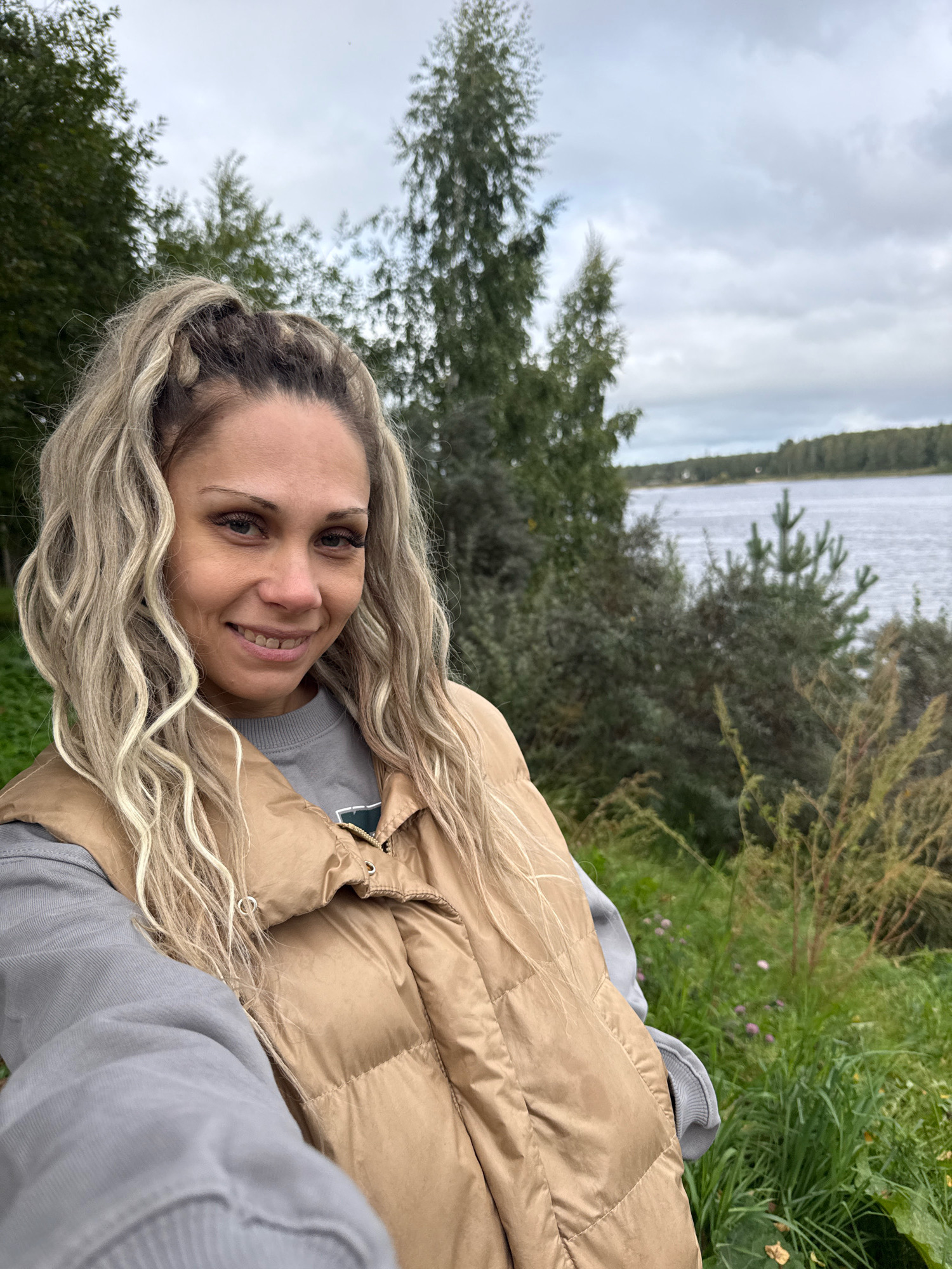 A medium-shot, selfie-style photograph captures a woman with blonde, wavy hair pulled into a high ponytail with braids framing her face. She is smiling and looking directly at the camera. She is wearing a grey sweatshirt underneath a tan, quilted vest. The background is a natural landscape with trees and a body of water, consistent with the location of Rusanovo, Russia. The sky is overcast with grey clouds. The foliage behind her appears dense and green, with some dry grass and small pink flowers in the foreground. The water in the background is grey and choppy.