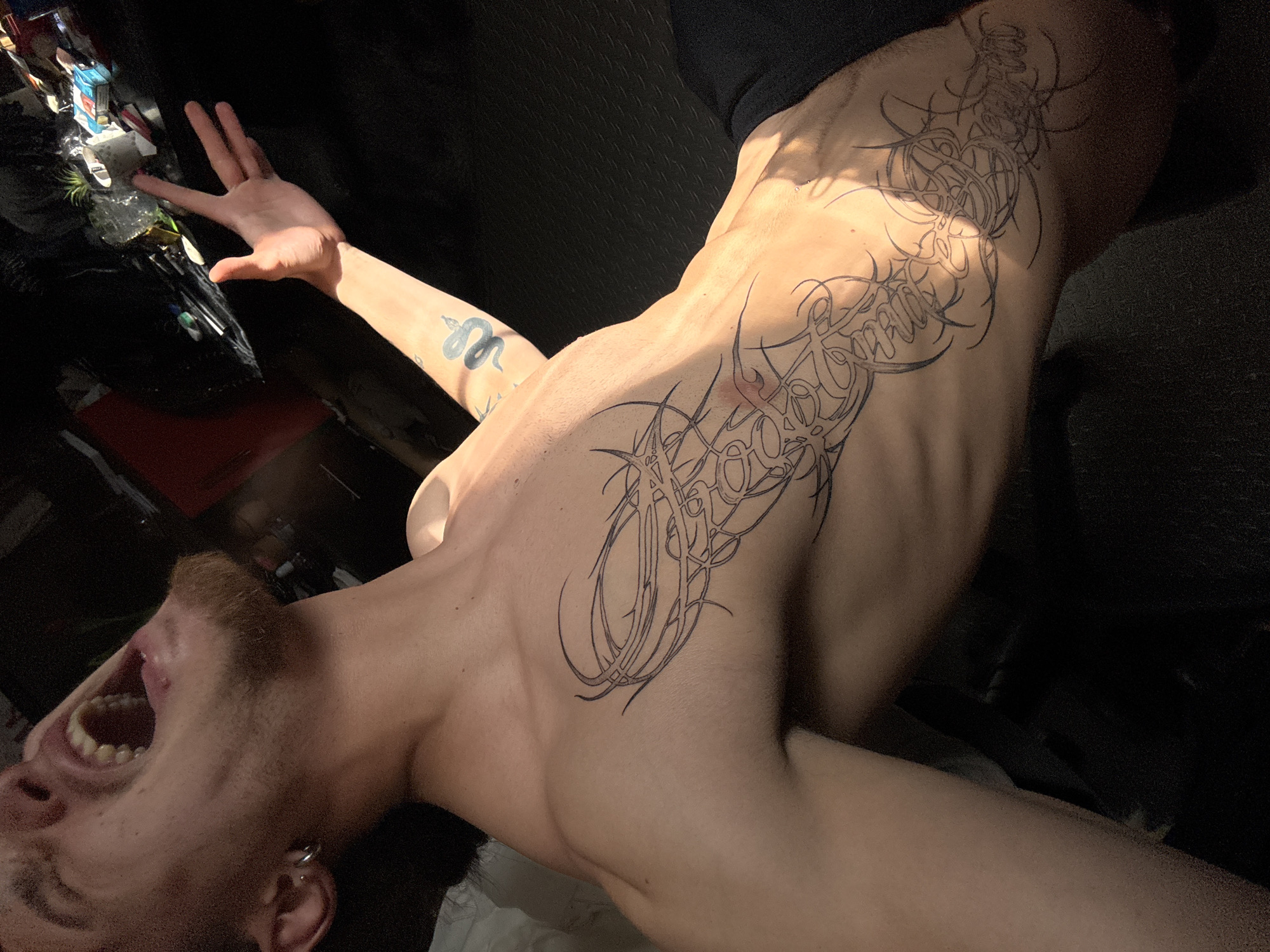 A shirtless man with a light beard and multiple tattoos is captured at a tilted angle. The tattoos, in black ink, feature abstract designs covering his torso and a snake motif on his left arm. He is tilting his head back, mouth open, possibly yelling. He has pierced ears. The background features indistinct, dark objects. The location of this image is unknown and there is nothing that indicates that it was taken in Sofia, Bulgaria.