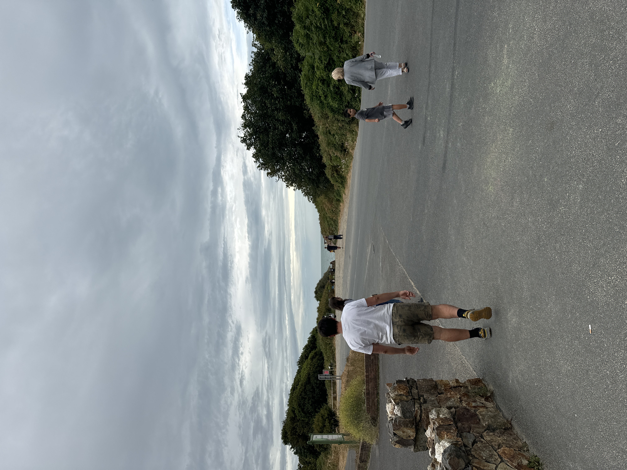 A group of people are walking on a paved path in Courtown, Ireland. The sky is overcast, with heavy clouds. There are trees and bushes on either side of the path. In the distance, the ocean can be seen. A man in a white t-shirt and patterned shorts walks away from the camera. To his right, a woman and a young boy walk towards the camera. Further in the distance, two other figures walk away from the camera. A low stone wall is visible on the left side of the path. A sign is also visible behind the stone wall.