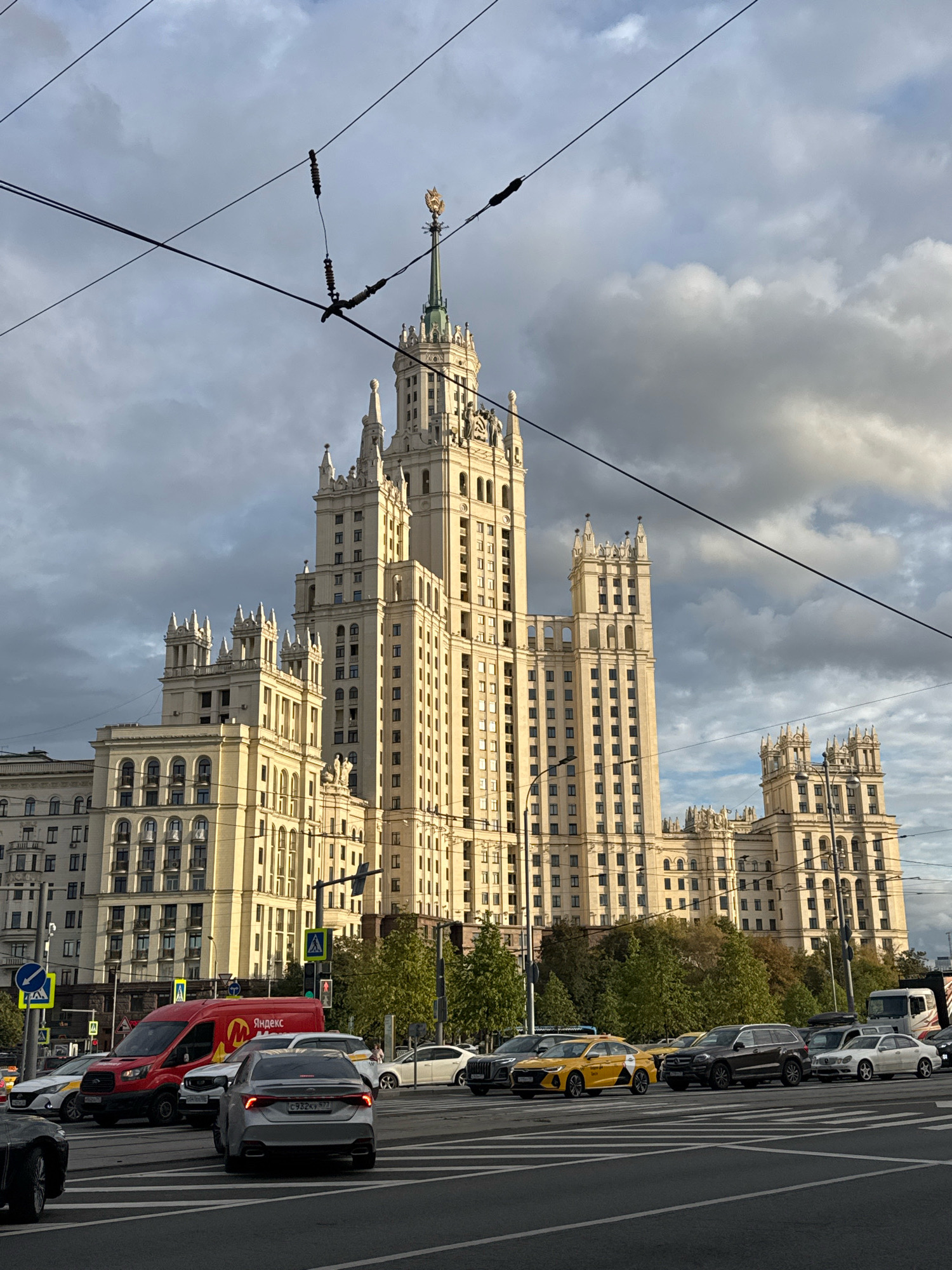 The image depicts a street scene in Moscow, Russia, featuring the iconic Stalinist-era skyscraper, Kudrinskaya Square Building, as the central architectural element. The sky is cloudy, with patches of sunlight illuminating the cream-colored building. The skyscraper is a multi-story structure with distinctive tiered spires, decorative elements, and numerous windows. In the foreground, a busy intersection is evident, with several cars on the road, including a red Yandex van, yellow taxis, and other passenger vehicles. Traffic lights and pedestrian crossing signs are visible. Overhead, trolley wires crisscross the sky, indicating public transportation routes. Trees line the background, and other buildings, also appearing to be of a similar architectural style, can be seen to the left and right of the main skyscraper. The overall impression is one of an urban environment with significant architectural heritage and active city life.
