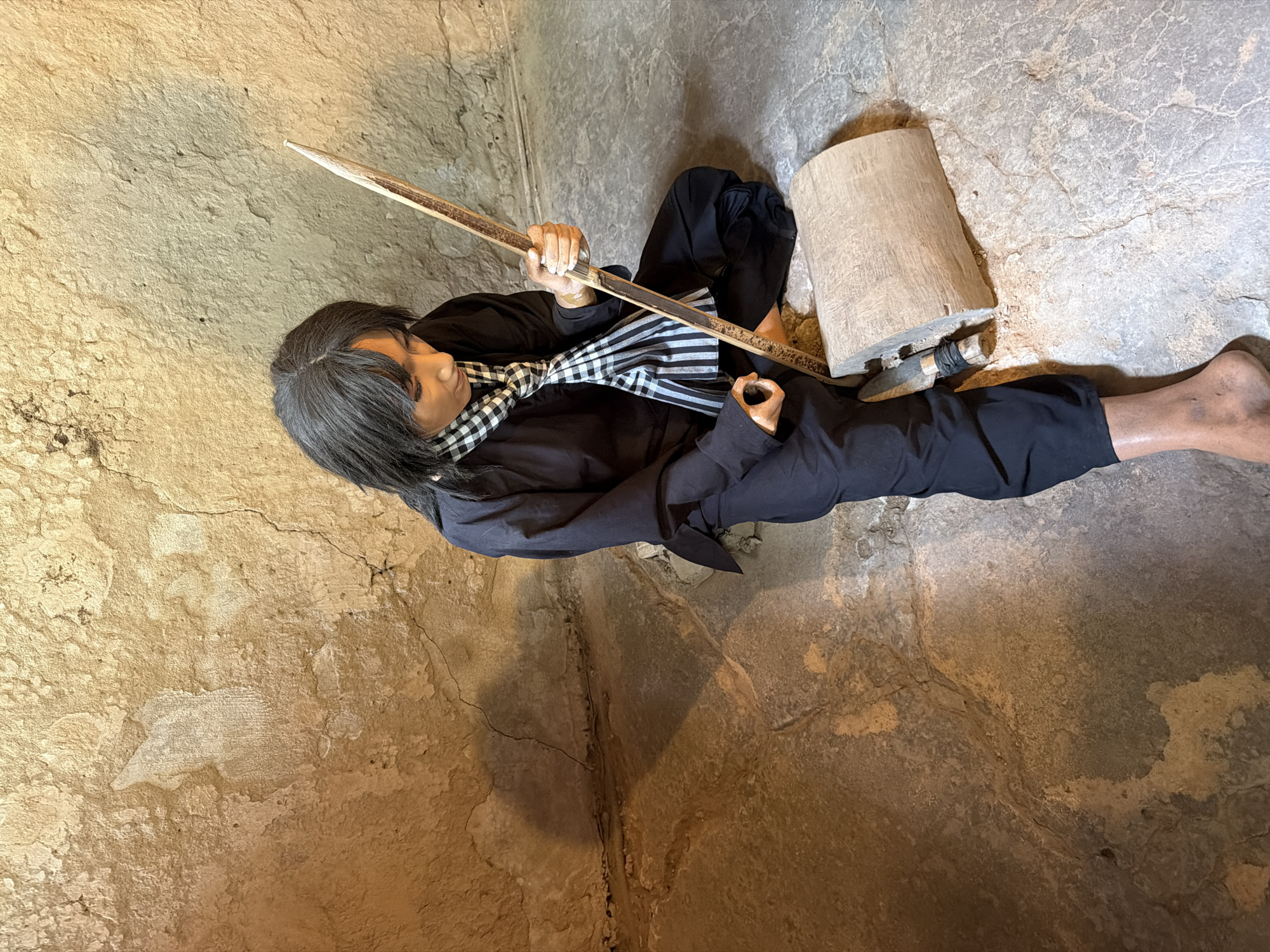 An overhead shot depicts a lifelike mannequin or statue of a person situated in what appears to be an underground tunnel or rustic dwelling, given the rough, cracked, and earthy textures of the ground and walls. The figure is clad in a black long-sleeved shirt, black pants, and a black-and-white checkered scarf, with dark hair cascading over their shoulders.
The person is positioned in a seated or crouched posture, holding a long, slender wooden stick with both hands, suggesting an activity involving crafting or using the stick as a tool. To their right, a roughly cylindrical piece of wood or log rests on the ground. A smaller tool, possibly a pickaxe or chisel with a wrapped handle, is partially embedded into this log.
The setting is dimly lit, emphasizing the rugged environment. The overall scene portrays an individual engaged in manual labor or a focused task, possibly wood carving or processing materials. The static nature of the figure, combined with the historical context of Ho Chi Minh City, Vietnam, suggests this is likely a diorama or exhibit, possibly depicting life or activities within the Cu Chi Tunnels or a similar historical site. No text is visible in the image.