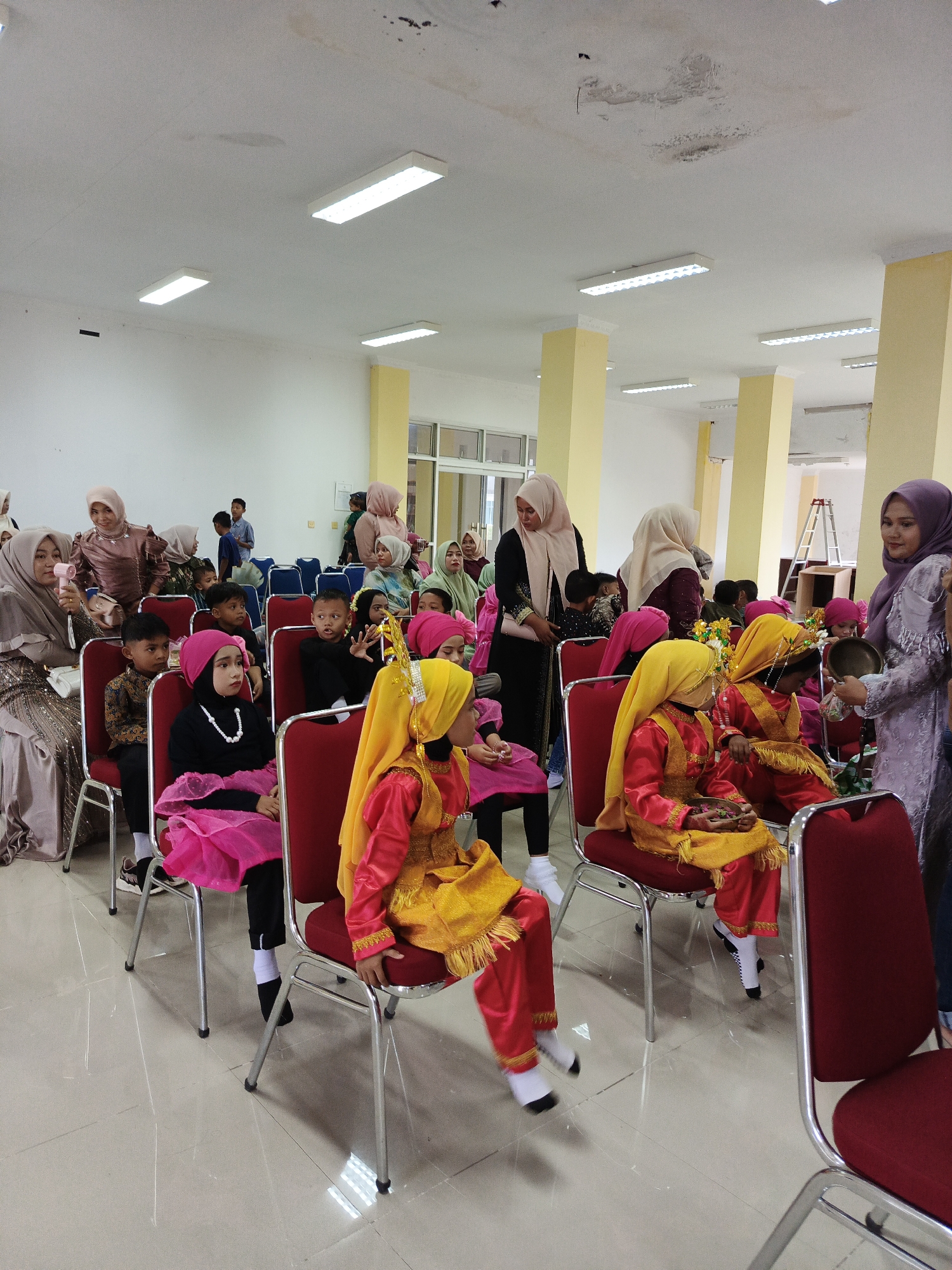 The image depicts an indoor gathering, likely a cultural performance or ceremony, taking place in Lhokseumawe, Indonesia. The scene is set in a well-lit room with a tiled floor and bright fluorescent lighting fixtures on the ceiling. Several rows of red-seated chairs with silver metal frames are arranged, occupied by a mix of adults and children.
In the foreground and midground, a group of children are seated, many wearing traditional attire. Some children are dressed in vibrant yellow and red outfits, complete with headscarves and ornate headwear, suggesting a performance. Others are in simpler, dark-colored clothing. The children appear to be watching something or waiting for their turn.
Adults are visible in the background, some standing and others seated. They are also predominantly wearing headscarves and modest clothing, consistent with cultural norms. One adult on the right is holding a small metallic object, possibly part of the ceremony. Another adult in the center appears to be addressing the children. The overall atmosphere suggests a community event with a focus on youth participation. The presence of yellow pillars in the background indicates a public building or event space.