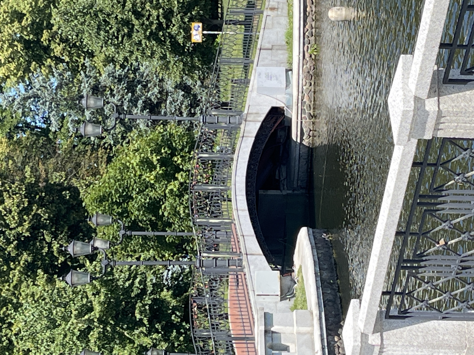 The image shows a canal with a stone embankment and a metal fence along its edge. A bridge spans the canal, with intricate metalwork visible on its railings. On the left side of the image, a lush green tree canopy dominates, with several vintage-style lampposts peeking through the foliage. A traffic light is visible in the background, with yellow and blue lights illuminated. The water in the canal is rippling, reflecting the sunlight.
The scene appears to be set in a park or a public area, likely in Kaliningrad, Russia, given the location context. The presence of lampposts and the bridge suggest a well-maintained urban or semi-urban environment. The time of day seems to be daytime, with bright sunlight casting shadows and illuminating the scene. The weather appears to be clear and pleasant, conducive to outdoor activities. No people are visible in the frame.
Notable details include the architectural style of the lampposts and the bridge's railing, which suggest a classic or historical design. The stone embankment and the rocky border of the canal add to the picturesque quality of the scene. The water has a slightly murky appearance, with some debris or natural elements floating on the surface. The green foliage of the trees provides a vibrant contrast to the stone and metal structures. There is a white, rectangular box-like object on the stone embankment near the bridge, the purpose of which is unclear.