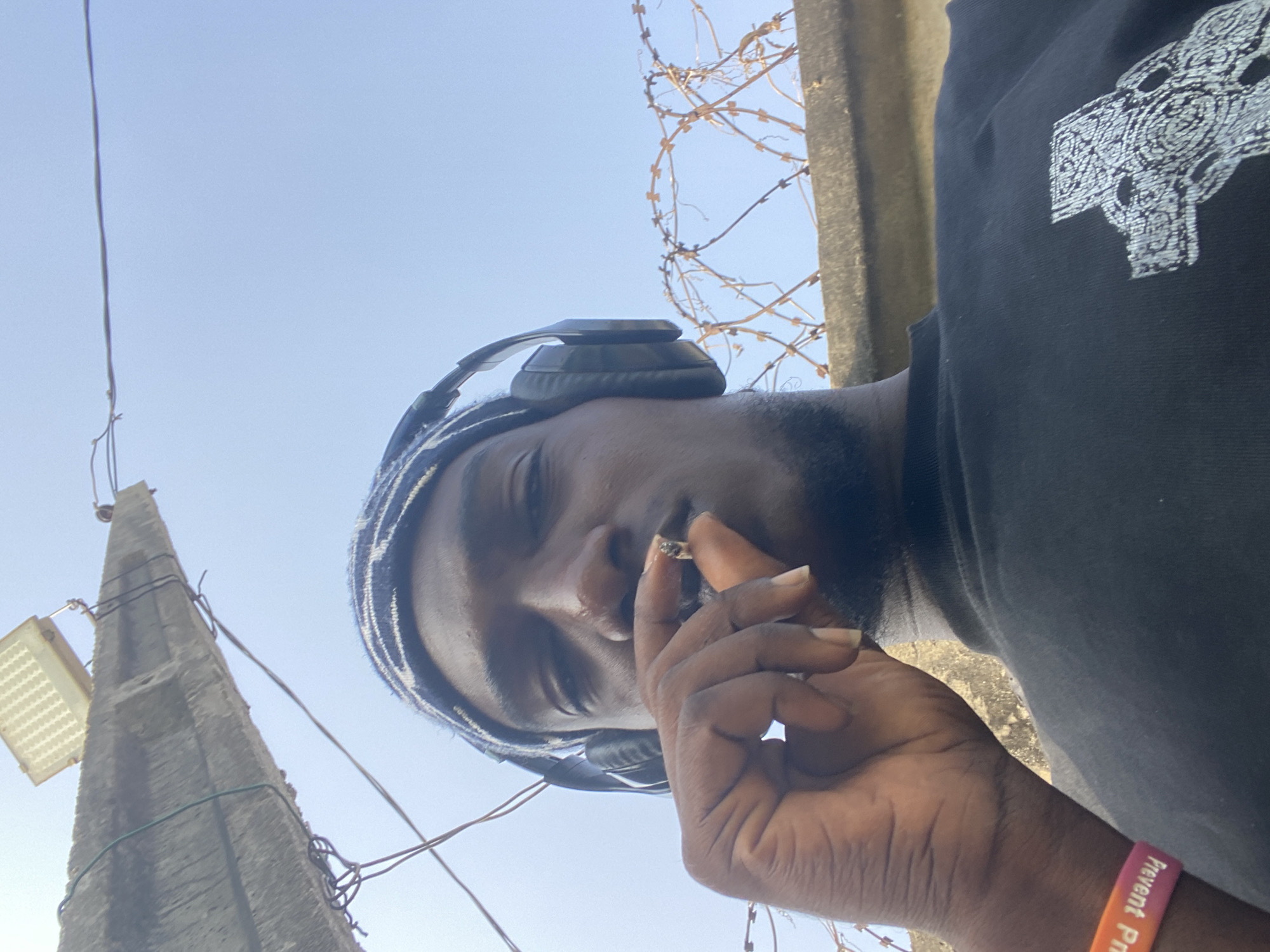 The image shows a dark-skinned man wearing headphones and a striped head covering, holding what appears to be a marijuana cigarette near his mouth. He is wearing a black T-shirt with a white design and a pink wristband that says "Prevent" partially visible. The background includes a utility pole with wires, a concrete structure topped with barbed wire, and a clear blue sky. The photo's angle suggests it was taken from a low perspective, looking upwards. Given the presence of utility poles and concrete structures, the location is likely an urban area. Without additional information or visual landmarks, it's impossible to pinpoint the exact location in Lagos, Nigeria.