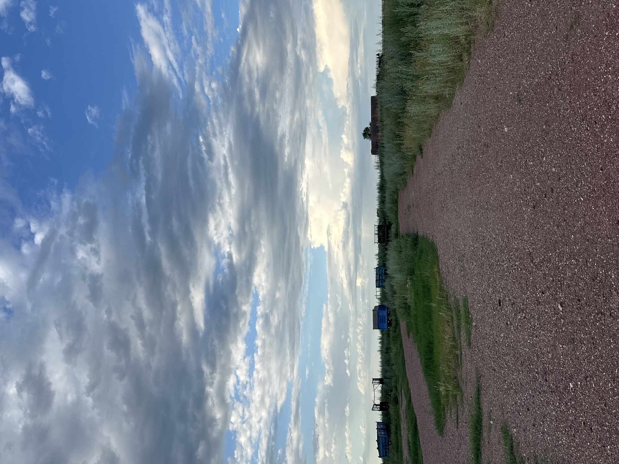 An outdoor, eye-level, vertical shot shows a gravel path leading into a field of tall green grass and scattered blue, boxy structures. The path is composed of small, reddish-brown pebbles. In the background, several blue, rectangular structures, some appearing to be old train cars or storage containers, are spaced throughout the field. A lone tree stands in the distance, near a low, rusty-brown building. The sky above is a dramatic mix of bright blue patches and thick, dark gray clouds. The overall impression is that of an abandoned or remote industrial site on the outskirts of a rural area, possibly in the region of Баягиз, Kazakhstan.