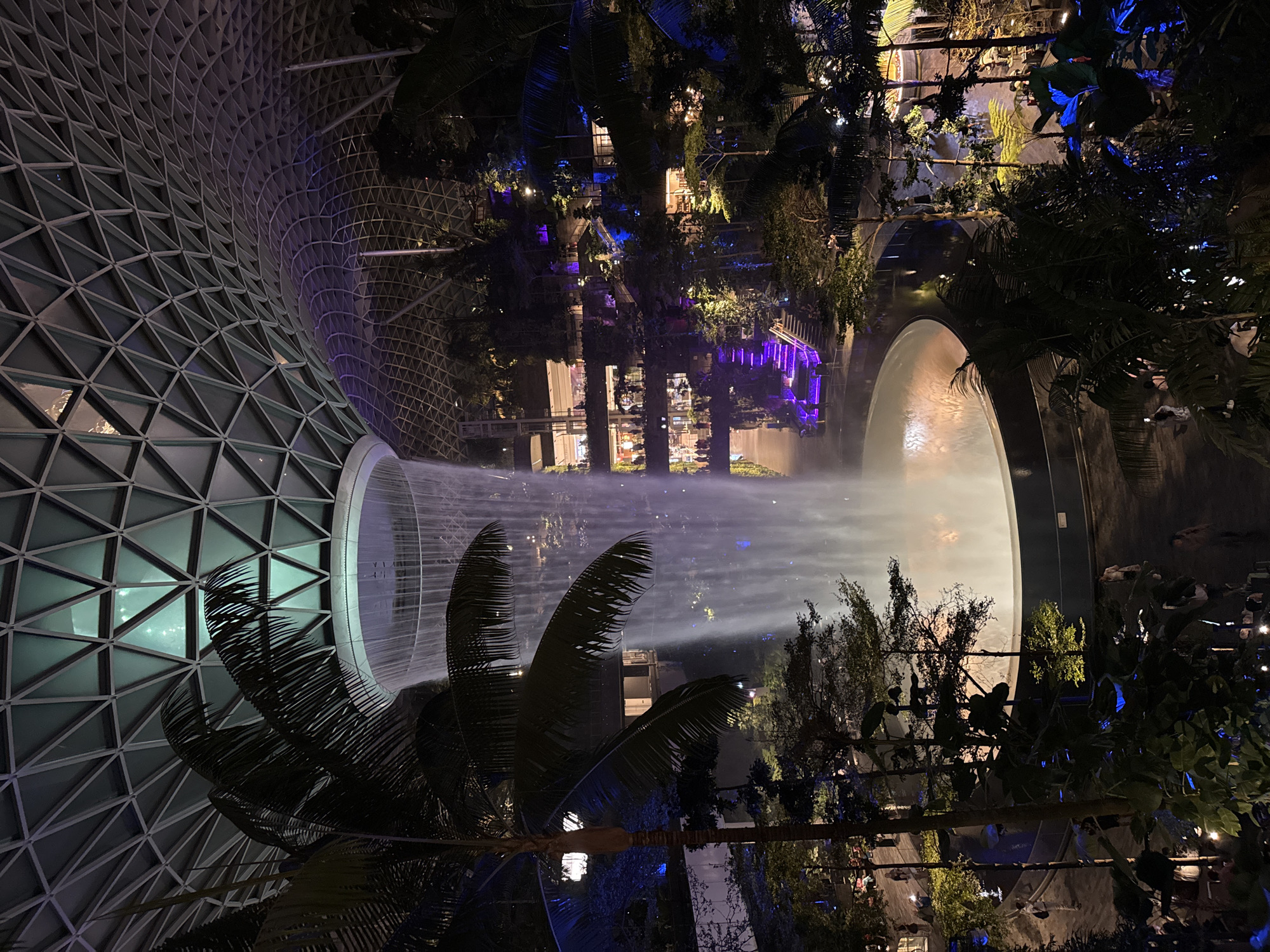 The image shows an indoor waterfall, the HSBC Rain Vortex, at Jewel Changi Airport in Singapore at night. The waterfall is a large, cylindrical structure in the center of the frame, with water cascading down from the ceiling into a circular pool below. The surrounding area is a lush, multi-level indoor garden with various trees and plants, illuminated by artificial lights in shades of blue and purple. Several levels of walkways and retail spaces are visible, with people walking and browsing. The ceiling is a geodesic dome made of glass and steel. Palm trees are visible in the foreground, framing the view. The overall atmosphere is one of a modern, futuristic, and vibrant urban oasis.